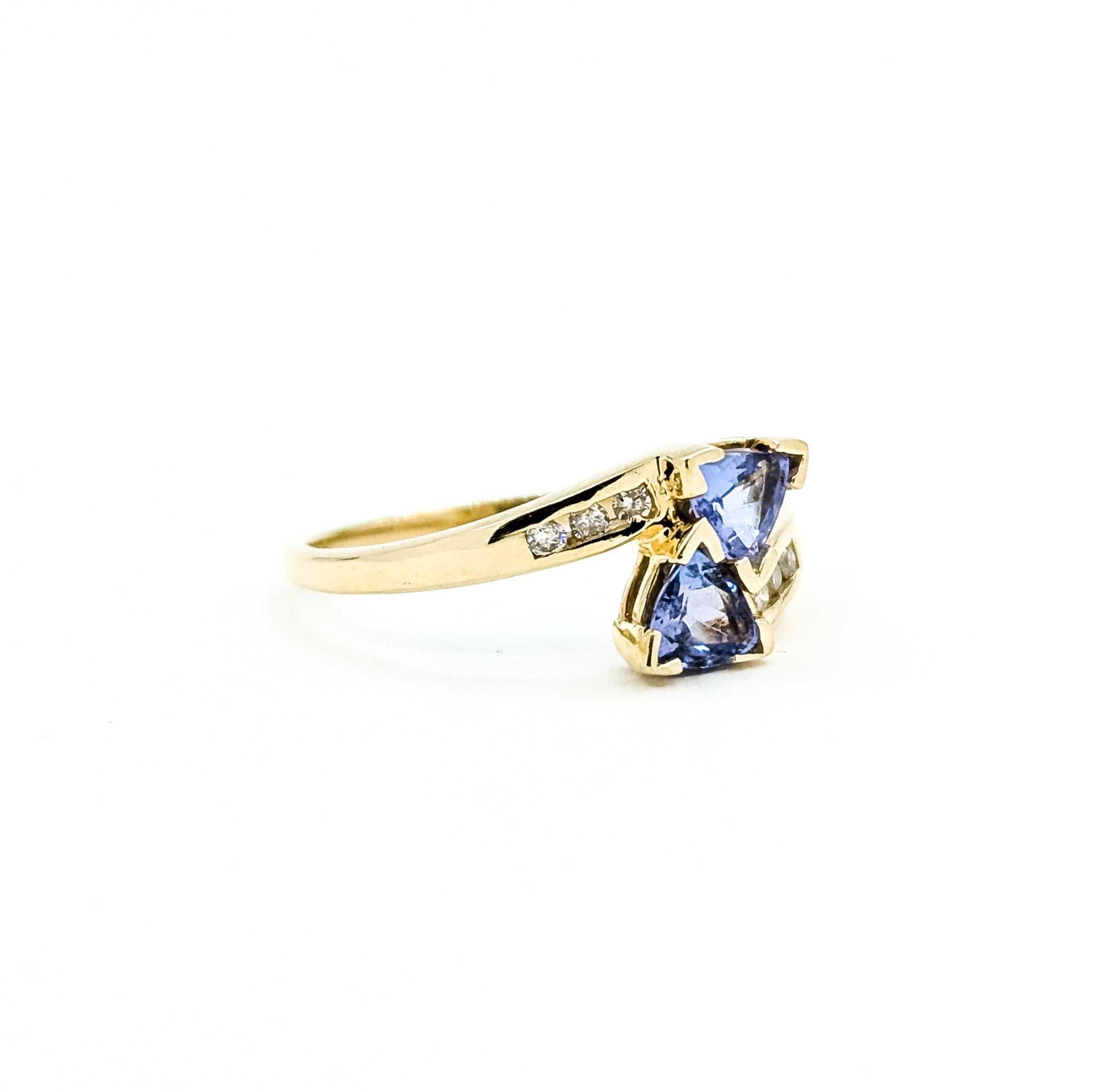 Cette bague gracieuse est réalisée en or jaune 14 carats et présente deux tanzanites de taille trillion serties à la main et d'une couleur bleu-violet vibrante. Des diamants ronds et brillants totalisant environ 0,06 carats, presque incolores et de