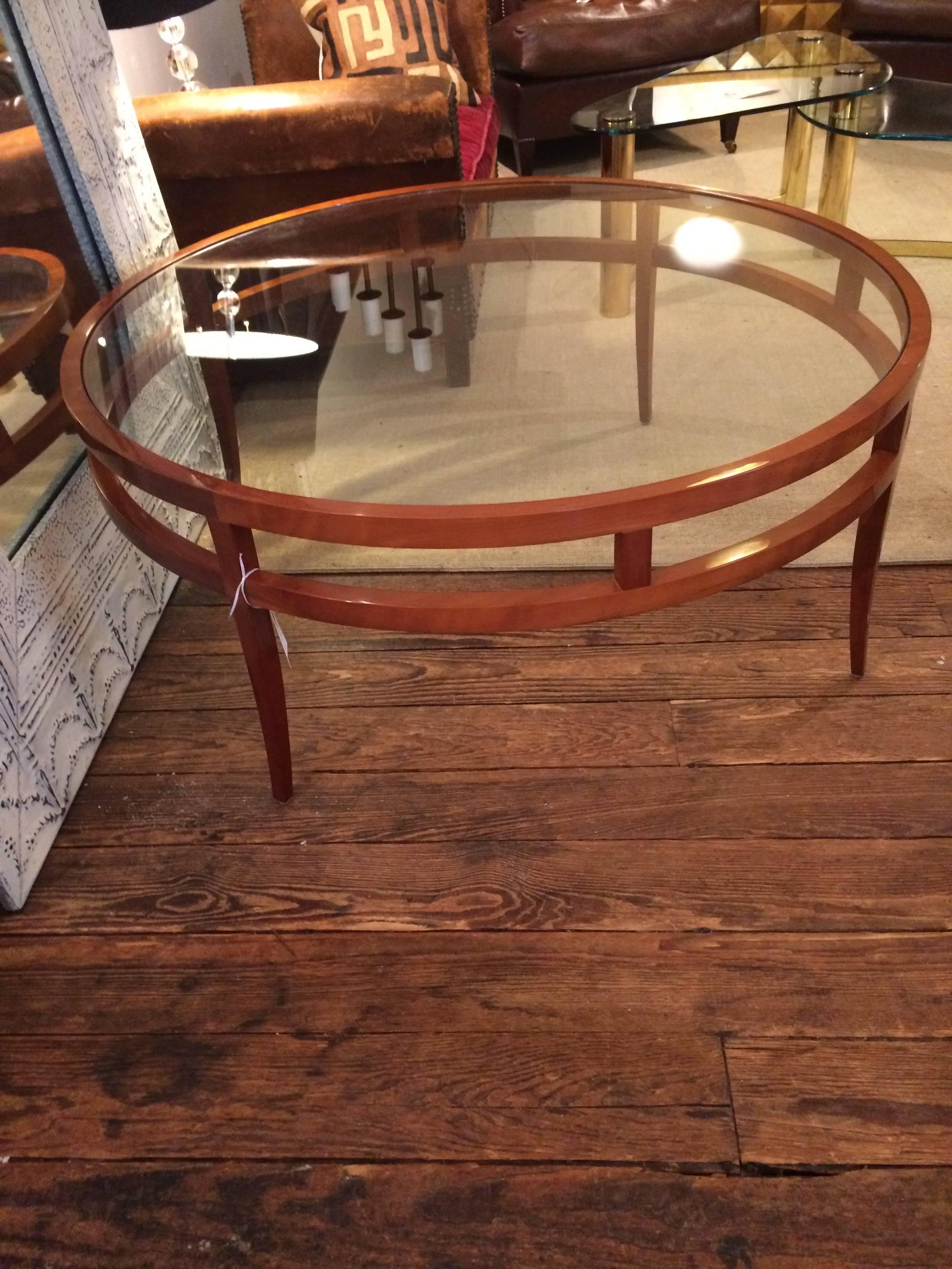 Sleek coffee table having a high gloss honey colored wooden cherry base with elegant tapered legs and a very large round glass top.