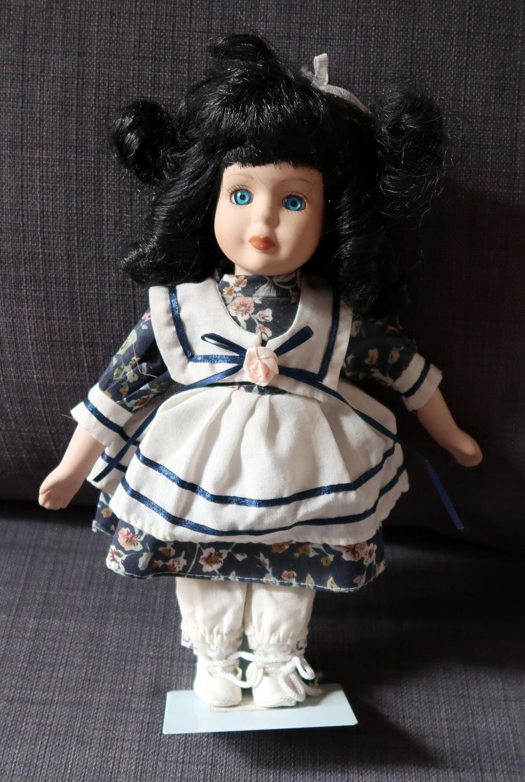 Immerse yourself in the charm of a bygone era with this Vintage Porcelain Doll, a delicate beauty hailing from the middle of the 20th century. Adorned in a white and blue dress, with striking black hair and captivating blue eyes, this doll is a