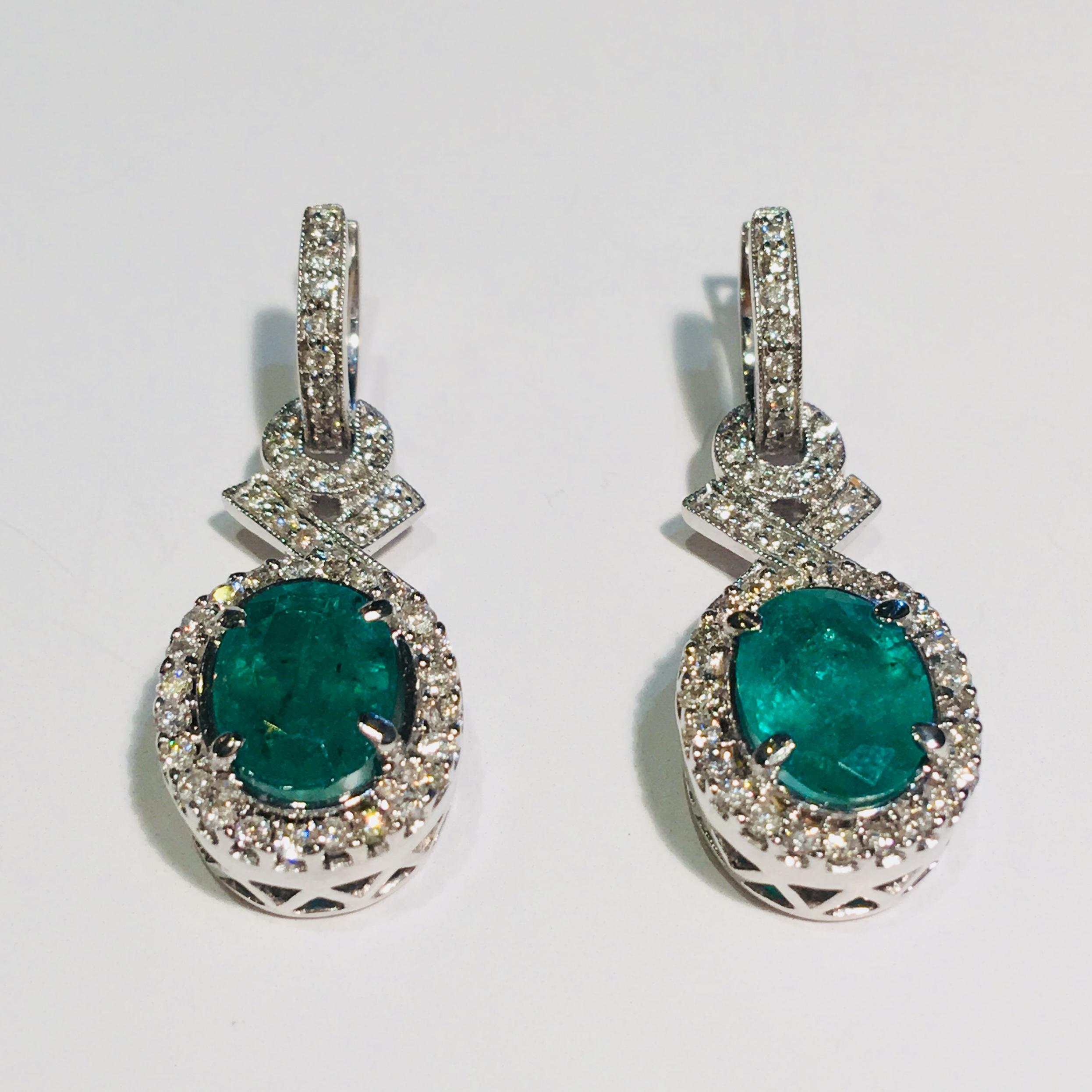 Elegant, dangling emerald and diamond halo 18 karat white gold estate earrings feature large, prong set, bright green, oval cut emeralds surrounded by sparkling halos of prong set round brilliant diamonds and accented by pave set round brilliant