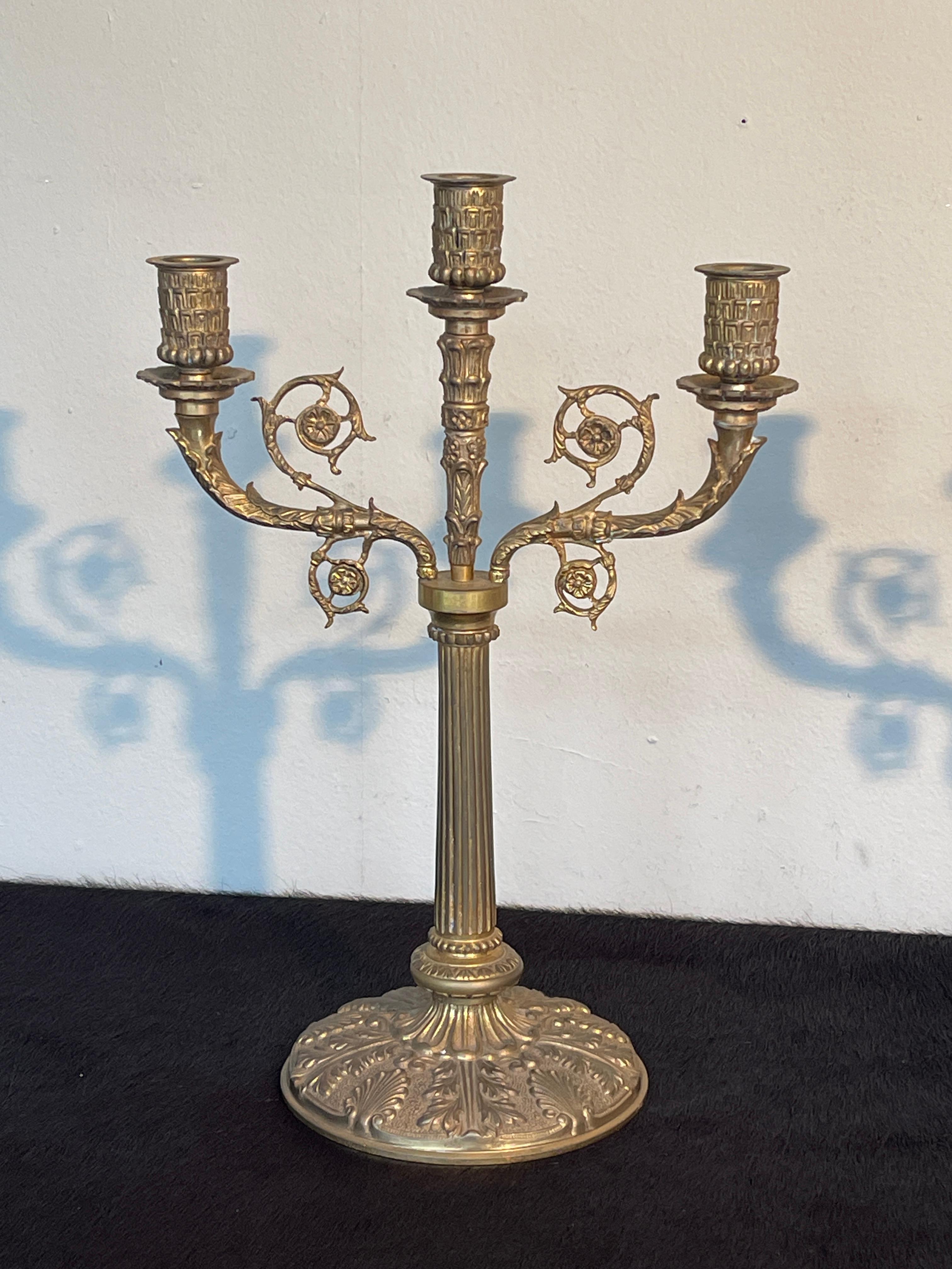 Magnifica coppia di candelabri a tre bracci in bronzo dorato, finemente lavorati con decori floreali e motivi ornamentali in stile classico. La base ampia e stabile presenta eleganti rilievi, mentre il fusto centrale scanalato sostiene con grazia i