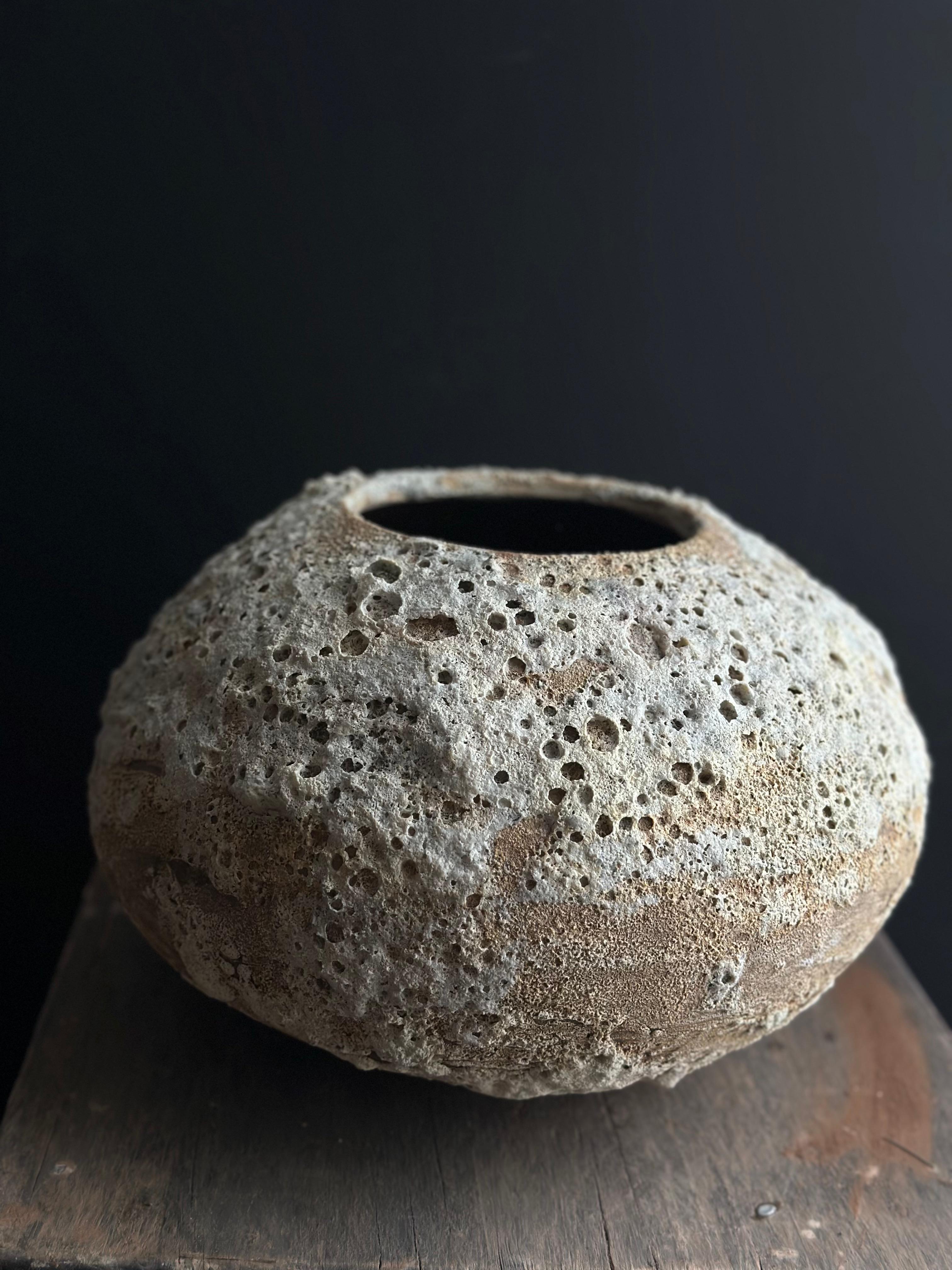 Questo vaso in ceramica fatto a mano ha una forma arrotondata e leggermente concava. La sua superficie è strutturata con numerosi piccoli fori e buchi irregolari, che le conferiscono un aspetto poroso e naturale. I toni del beige chiaro e del taupe