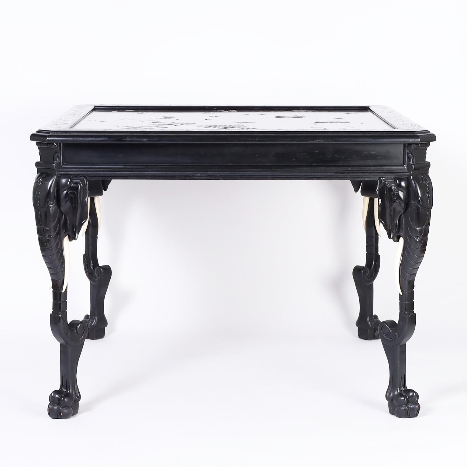 Square center table with a japanned or ebonized finish featuring a lacquered panel on top with an embossed stylized landscape. The legs have carved elephant heads with bone tusks and paw feet.