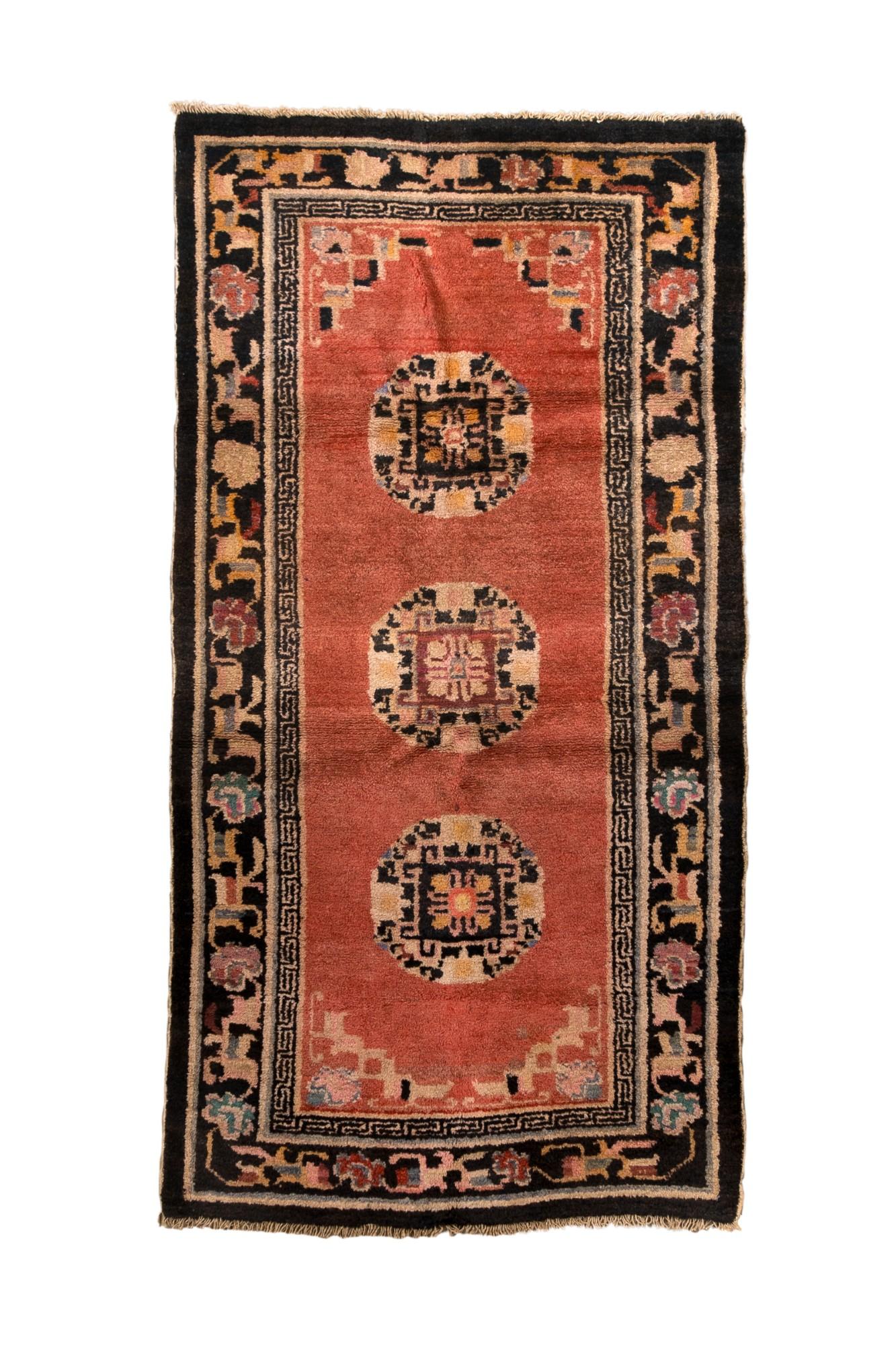 The open red-brown field presents three identical round medallions, with floral wreaths surrounding central square fretworks. Stepped fretwork corners. Black border with a paeony, leaf and palmette scroll. Greek key fretwork inner border. Thick