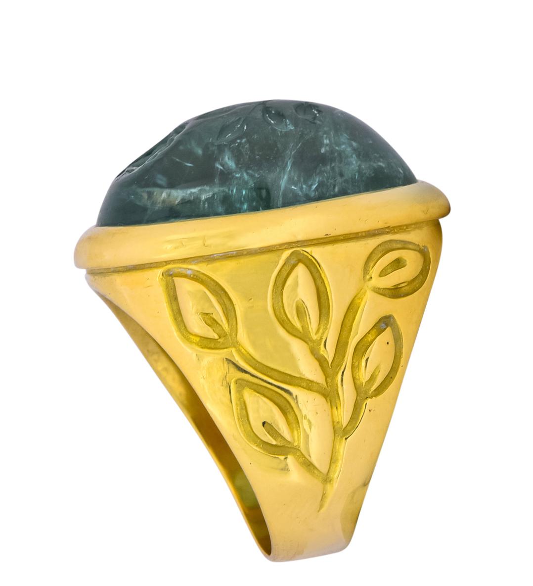 Centering an oval cabochon aquamarine deeply carved depicting a stemmed rose, measuring approximately 1 x 7/8 inch

Aquamarine is bezel set in a polished gold surround, semi-transparent and medium-dark greenish-blue in color

With deeply engraved