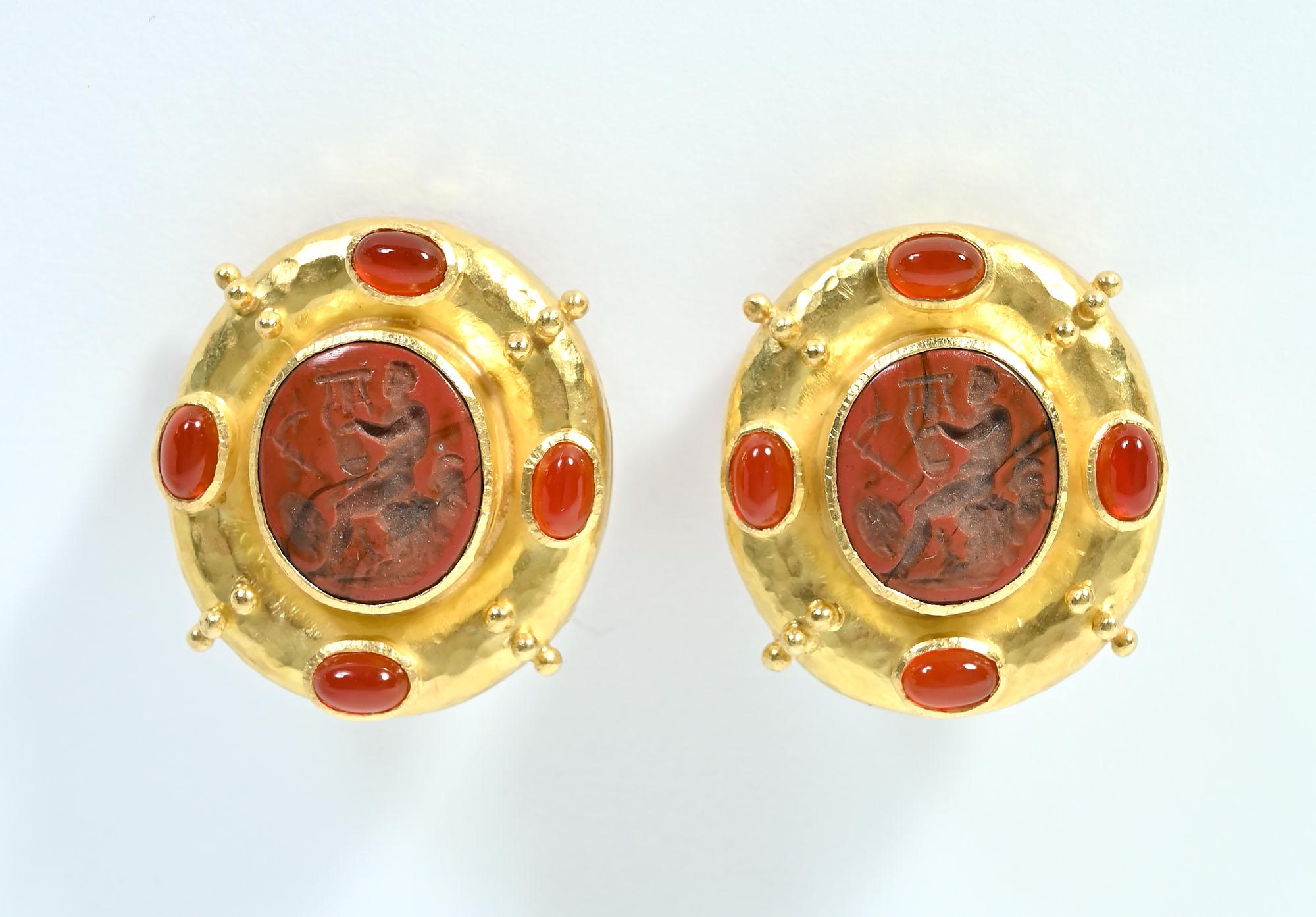 Large, bold earrings by Elizabeth  Locke. The central oval of jasper depicts a man in profile playing a musical instrument, Around the center medallion are four cabochon carnelians, Four groups of small gold balls are set at an angle between the