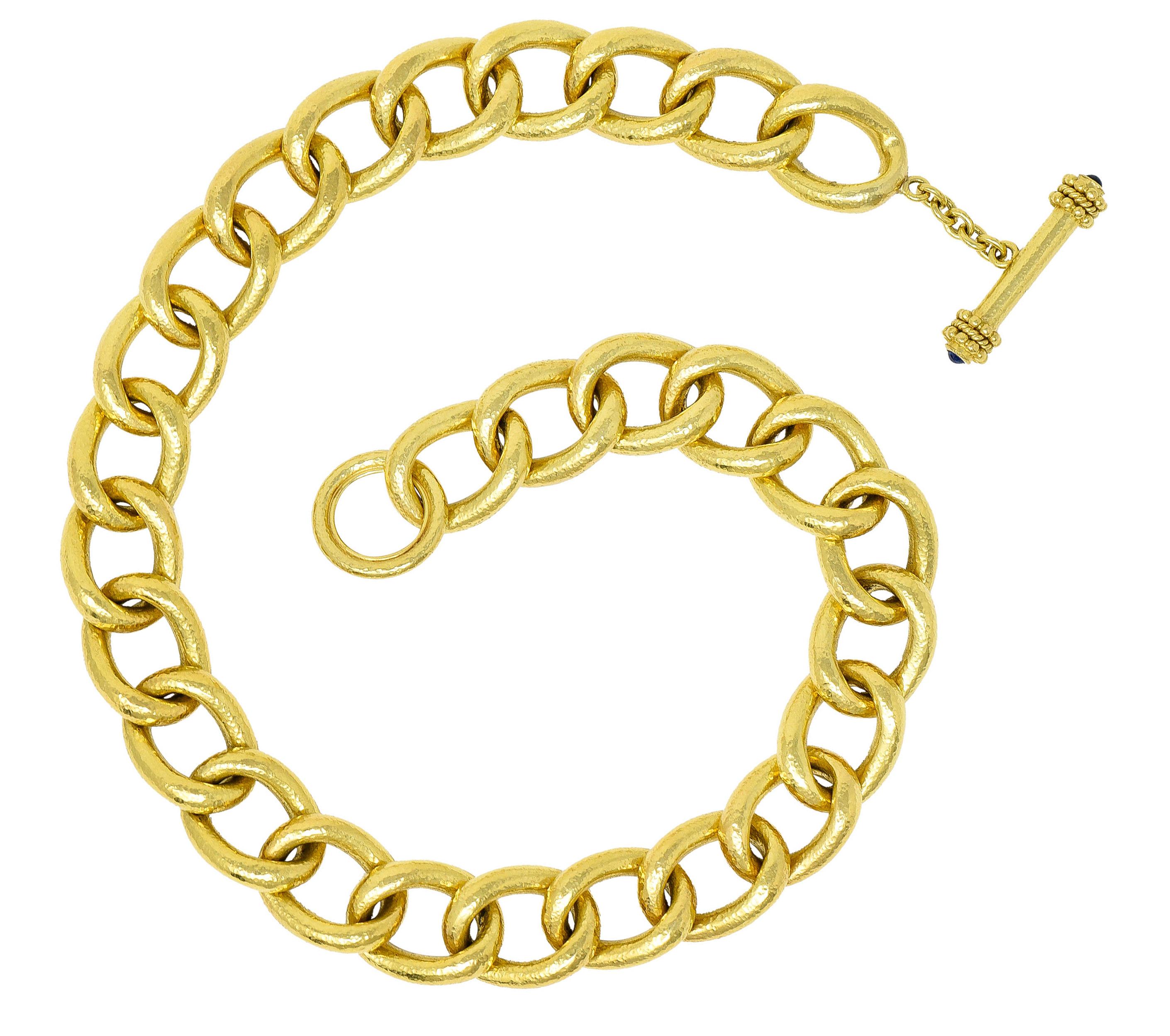 Collar necklace is comprised of large curbed links with a hammered finish

Completed by a bar toggle clasp with a twisted rope motif and gold bead detail

Bar terminates at each end as a bezel set 3.0 mm round sapphire cabochon - vibrantly