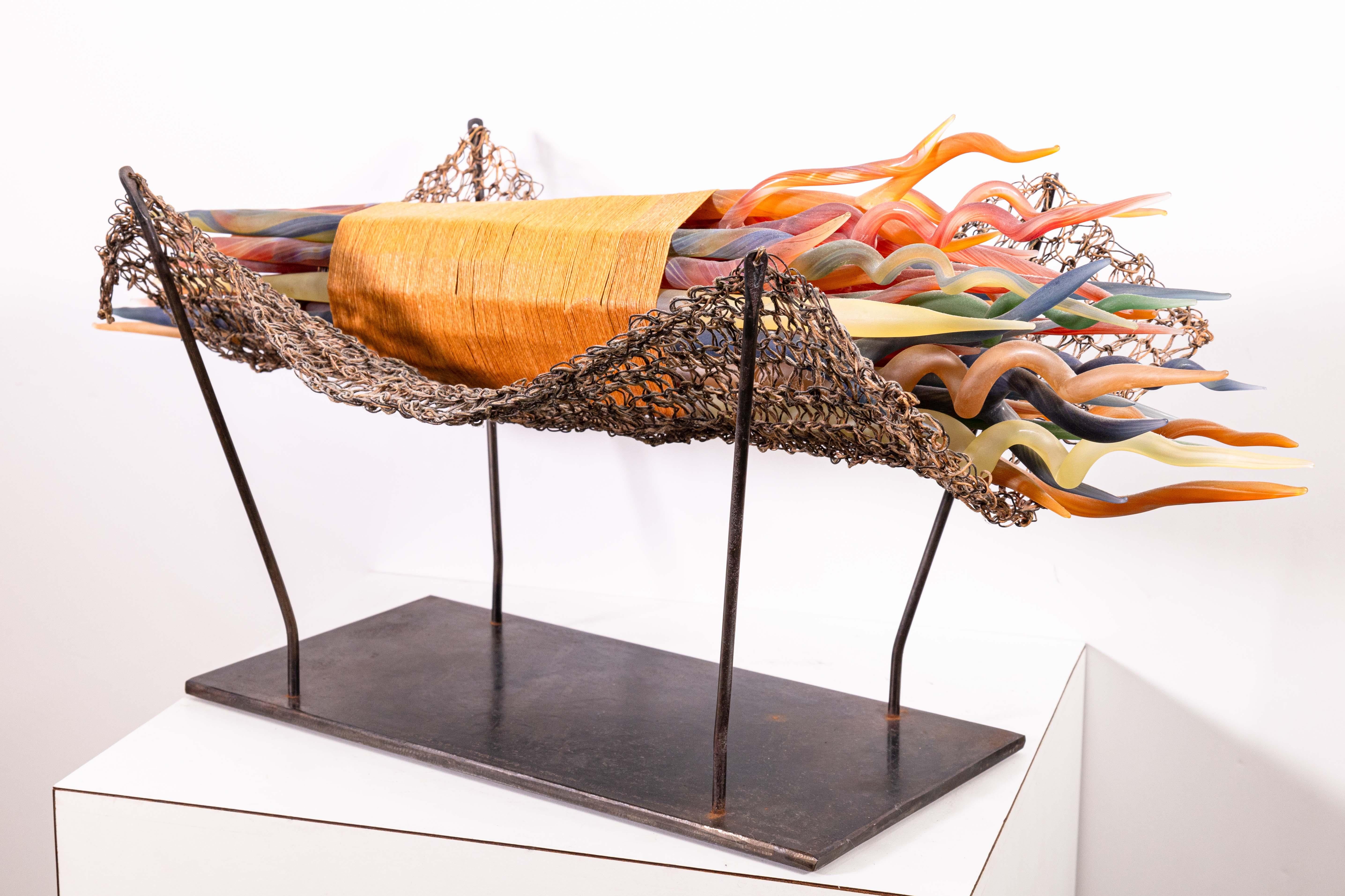 A contemporary glass sculpture by Elizabeth Ryland Mears. Circa 1990s. Small multi-colored glass sculptures are forged in the shape of twigs. They are spindled together, placed on a metal woven cradle, and suspended on a metal base.  Contemporary