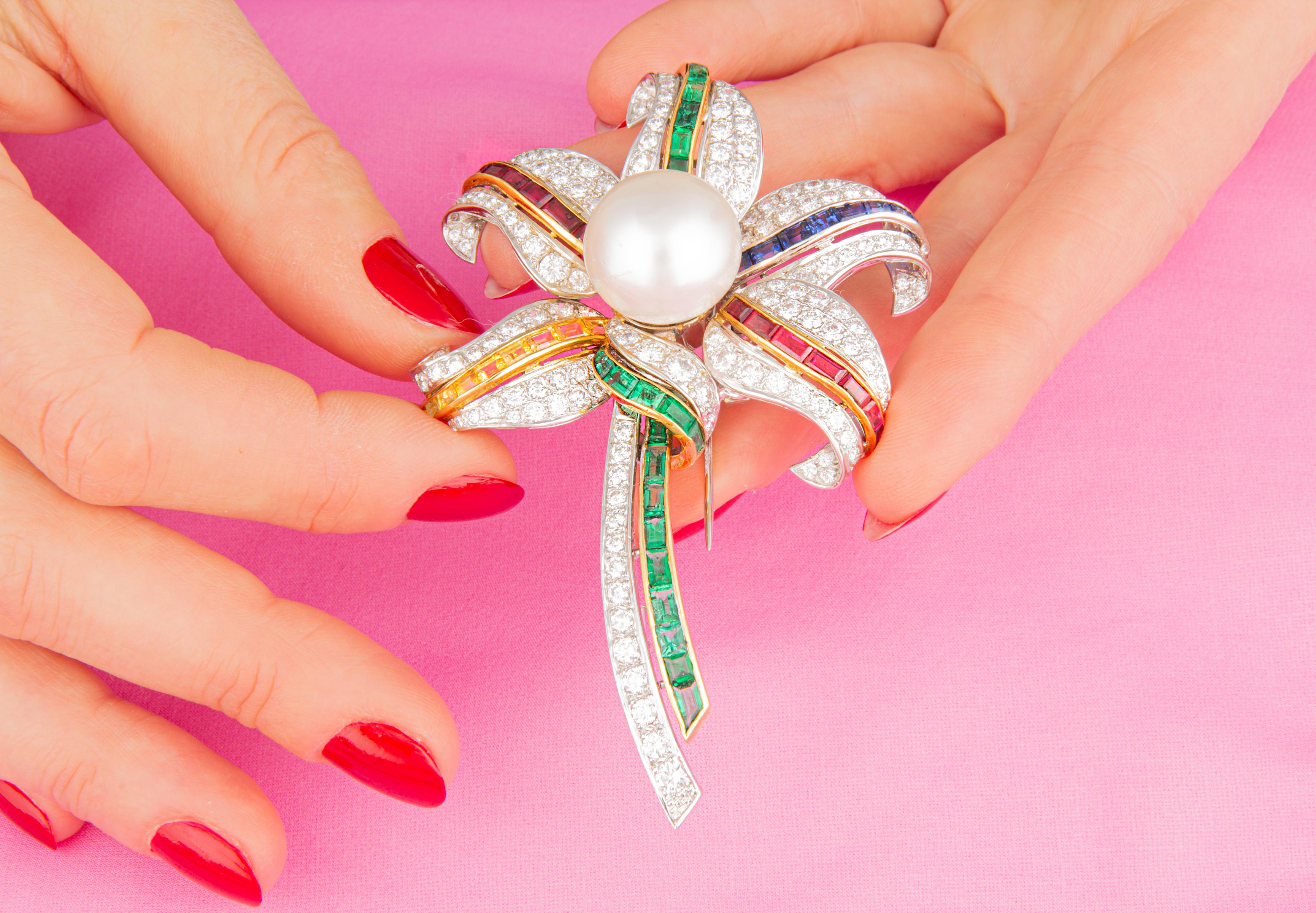 The flower diamond brooch features five leaves or petals around a large and lustrous South Sea pearl. The leaves and stem are each accented by a ribbon of custom cut baguette shaped precious stones of fiery color: Emerald (3.40 carats), ruby (3.57