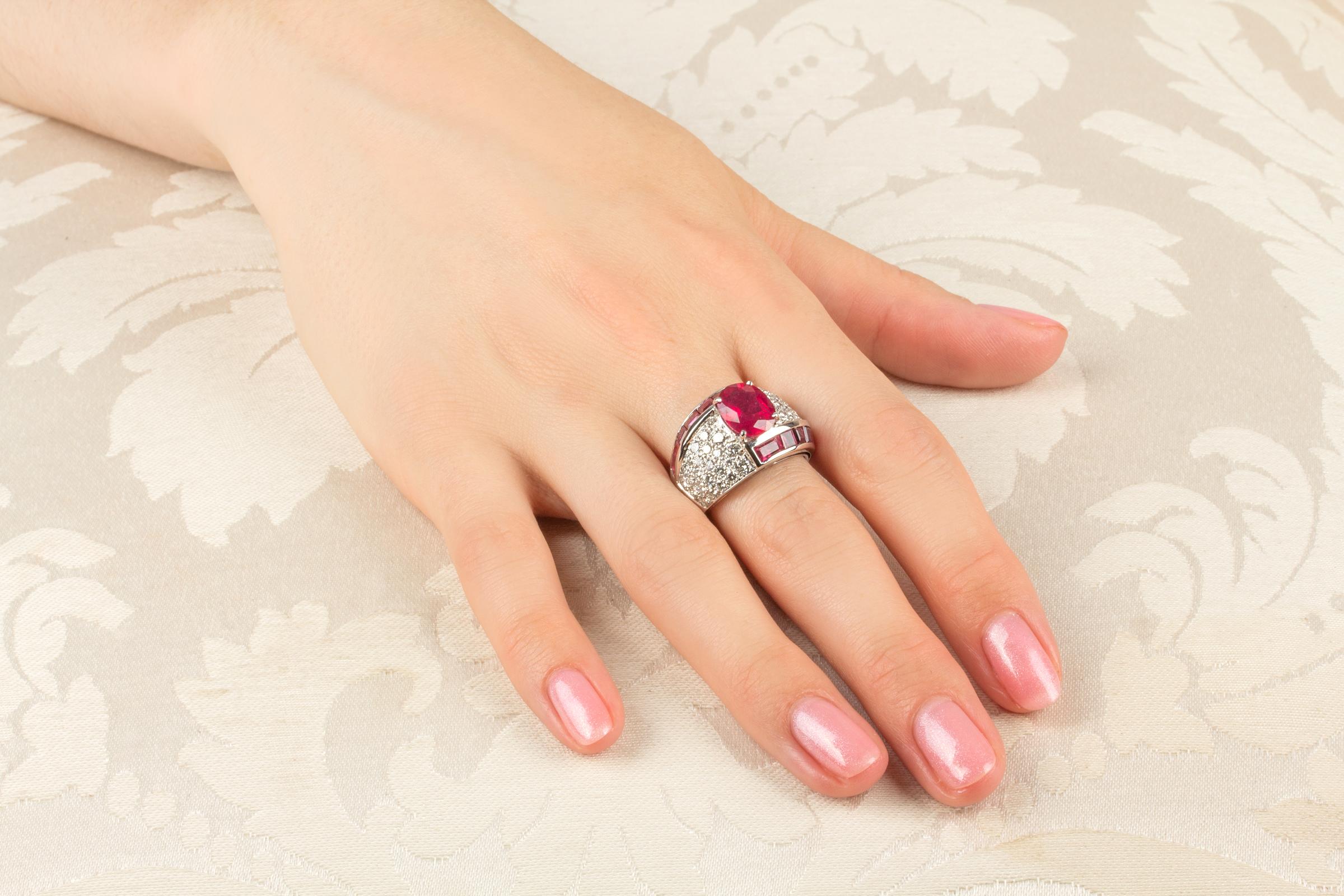 The ruby and diamond cocktail ring features an oval cut faceted  ruby weighing approximately 3 carats. The center stone is flanked by 2 ribbons set with custom cut channel set rubies for a total weight of 2.50 carats. The design is complete with