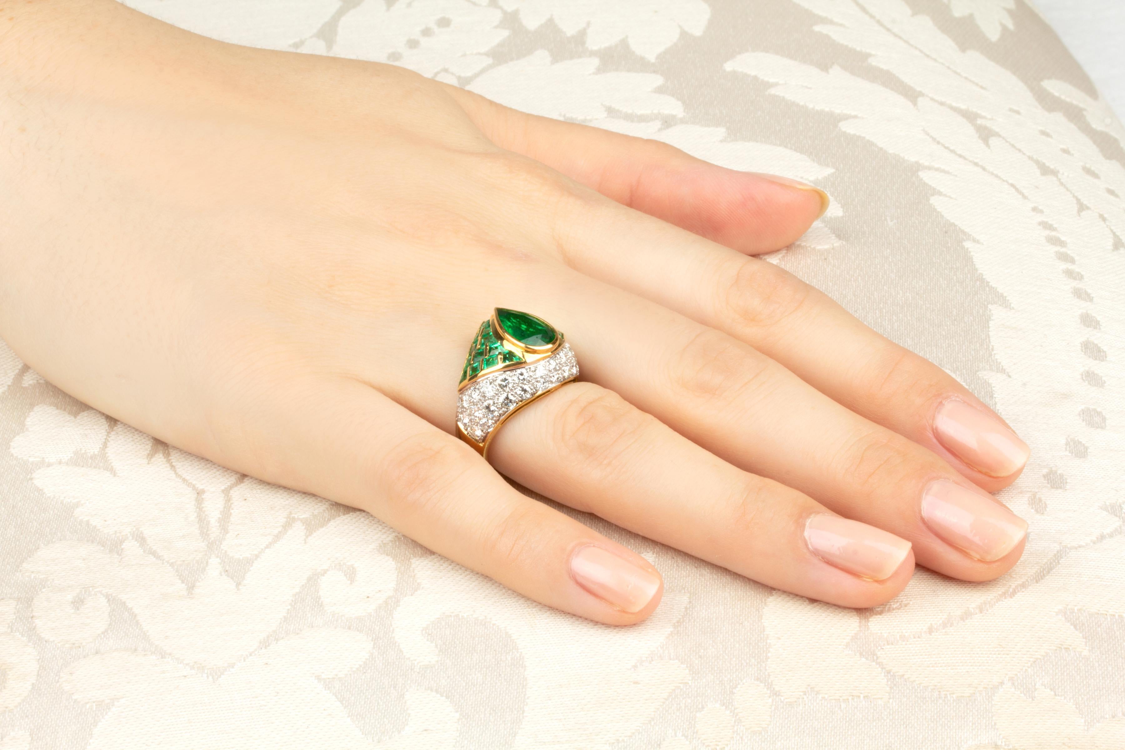The ring features a drop shape faceted emerald of brilliant color (1.64 carats) flanked by a geometric pattern of custom cut square emeralds (2.43 carats). The design is complete with 1.75 carats of round diamonds of top quality set en pavé