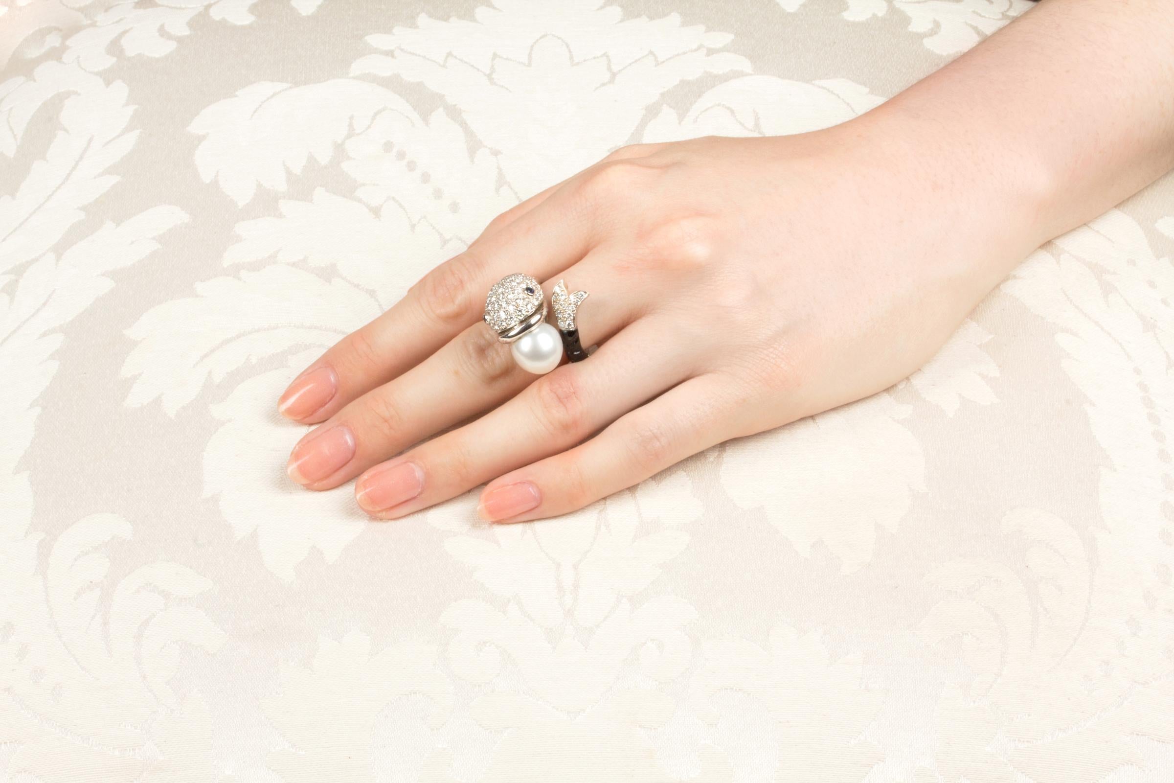 The fish ring with diamonds features a fish holding a  South Sea pearl of 11.5mm diameter in its mouth. The pearl is untreated. It displays a fine nacre and its natural color and luster have not been enhanced in any way. The diamonds decorating the