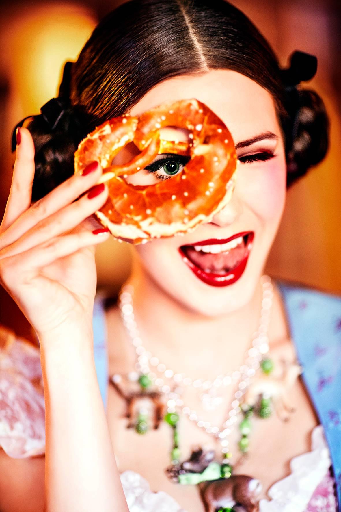 Color Photograph di Ellen von Unwerth - Brezel - Ragazza in Dirndl che guarda attraverso un Brezel, Fotografia Fine Arts, 2015