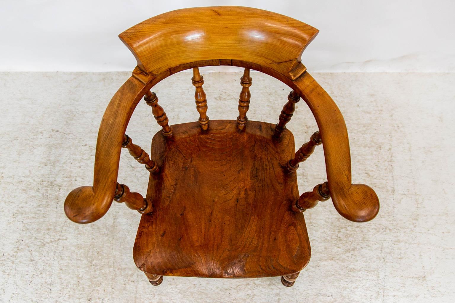 This captain's chair has scrolled arms and a flared back rest. The arms are supported by four turned spindles. The elm seat has a slight saddle shaping. The four turned legs are connected by a turned 
