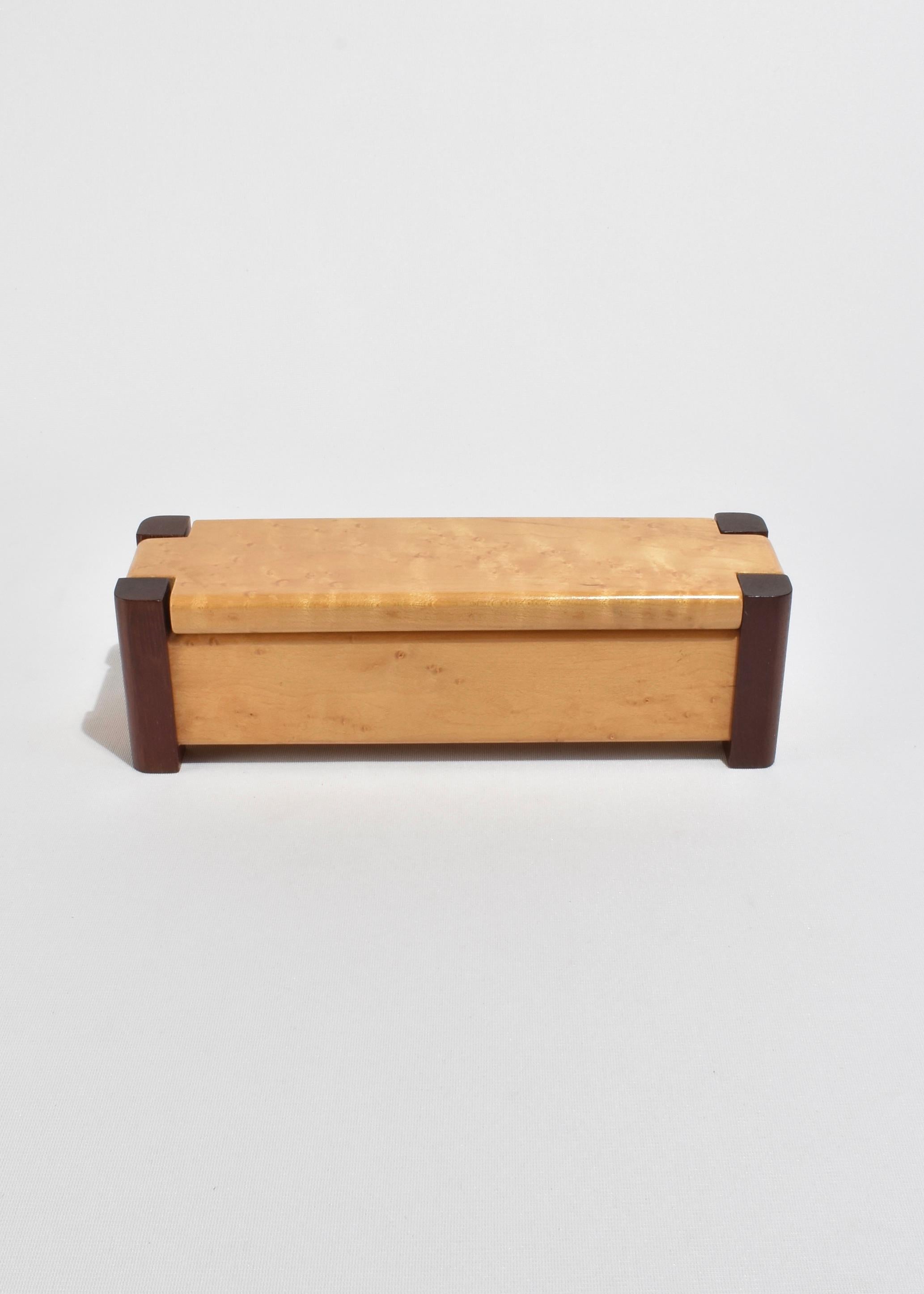 Una caja rectangular con tapa, hecha a mano en arce ojo de pájaro con juntas de esquina de madera oscura en contraste. La tapa encaja limpiamente en la parte superior, revelando un interior forrado de fieltro ideal para joyas o pequeños recuerdos.