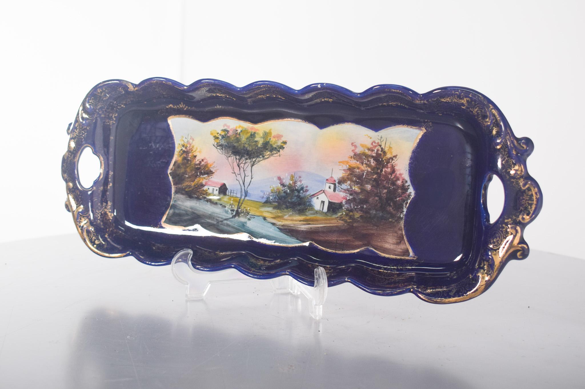 Sartén de barro ELPA (Elias y Paiva) de Alcobaça. Decorada en azul oscuro y dorado a lo largo de los bordes ondulados y festoneados. En el centro de la pieza, una pintura delicada y detallada, que representa un paisaje campestre con una iglesia de