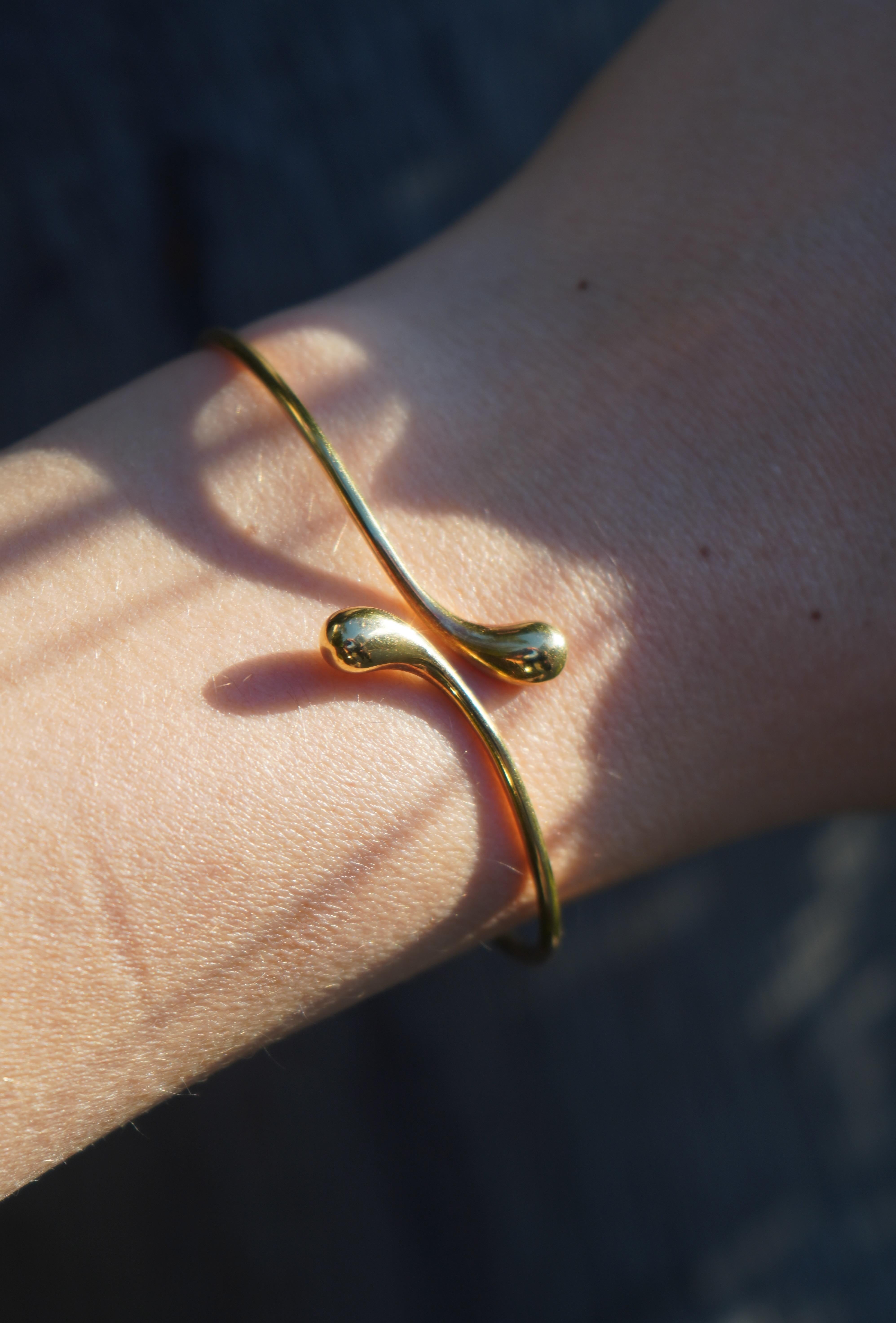 This Elsa Peretti for Tiffany & Co. 18k Gold Teardrop Bypass Bangle Bracelet features a sleek and minimalist design. The open-ended bangle curves gracefully, with each end forming a polished teardrop shape. Measuring 7