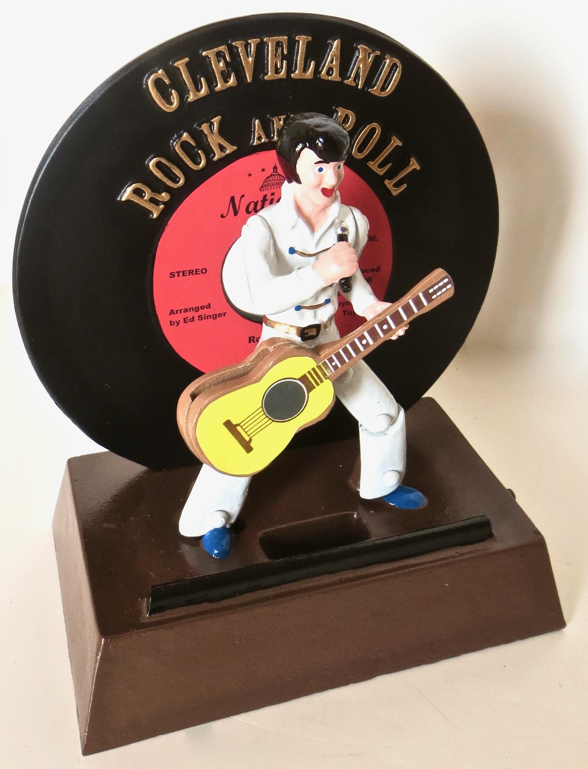 Made to commemorate the Rock and Roll Hall of Fame located in Cleveland, Ohio, this cast aluminum mechanical bank depicts Elvis Presley in full gala attire, typical of Elvis, with white suit, microphone in hand, and guitar fronting his rock and roll