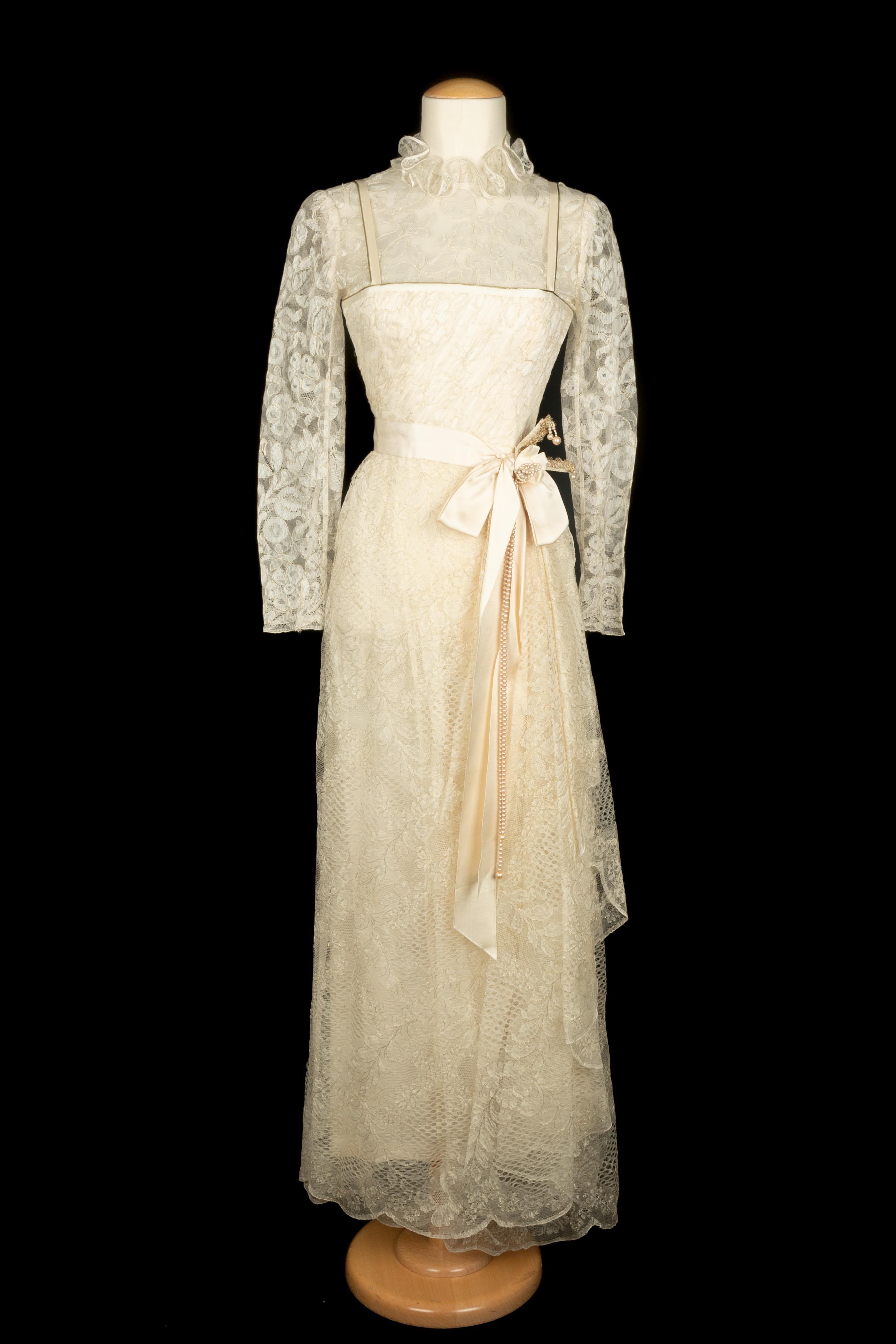 UNGARO - (Made in France) Robe de soirée en dentelle mécanique ivoire avec des fils dorés. Col à volants et manches longues. Ceinture en satin ornée de perles et d'un nœud. Étiquettes de composition et de taille manquantes, il convient à un 36FR.