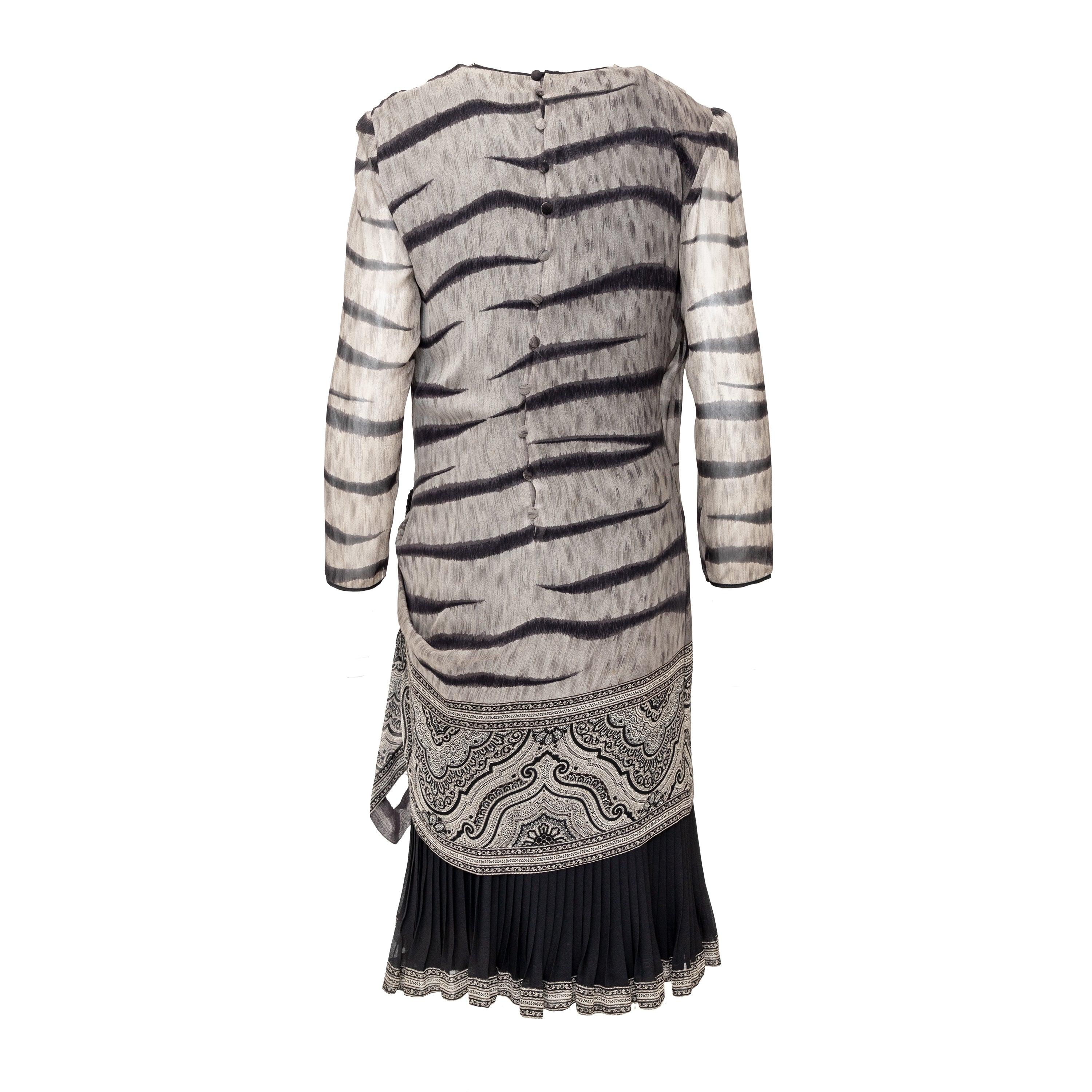 Presentamos el Vestido de Seda Ematite. Confeccionado en suave gasa de seda y de estilo envolvente, este vestido luce un elegante estampado animal en gris y negro con un bonito diseño paisley en el bajo. La parte inferior presenta pliegues de