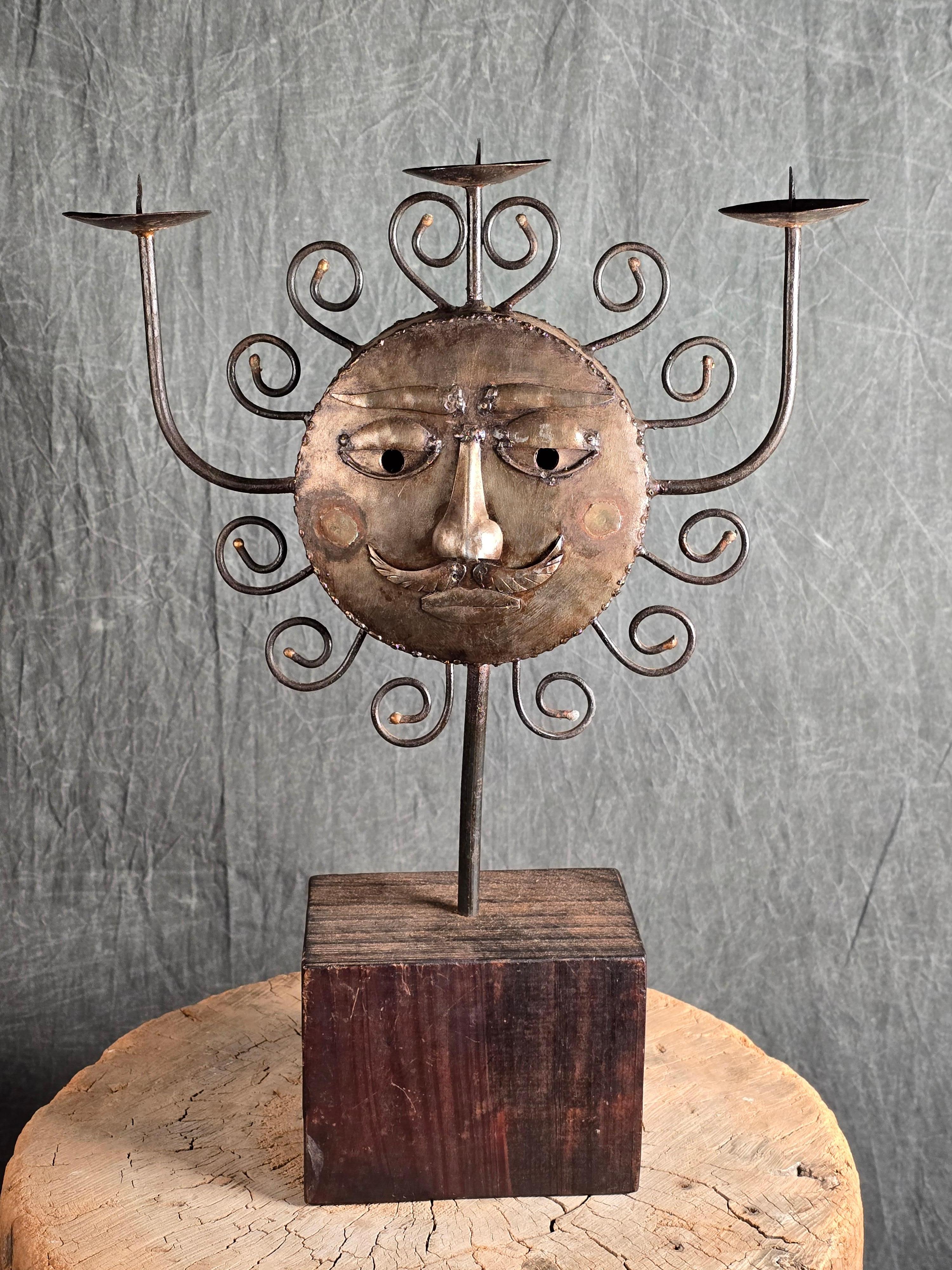 Candelabro in ferro forgiato a mano dal laboratorio benedettino Emaús, Messico, anni '60. Scolpito a forma di sole stilizzato con un volto umano su un lato e una mezzaluna sul retro, incorniciato da volute a raggiera. Il pezzo contiene tre candele
