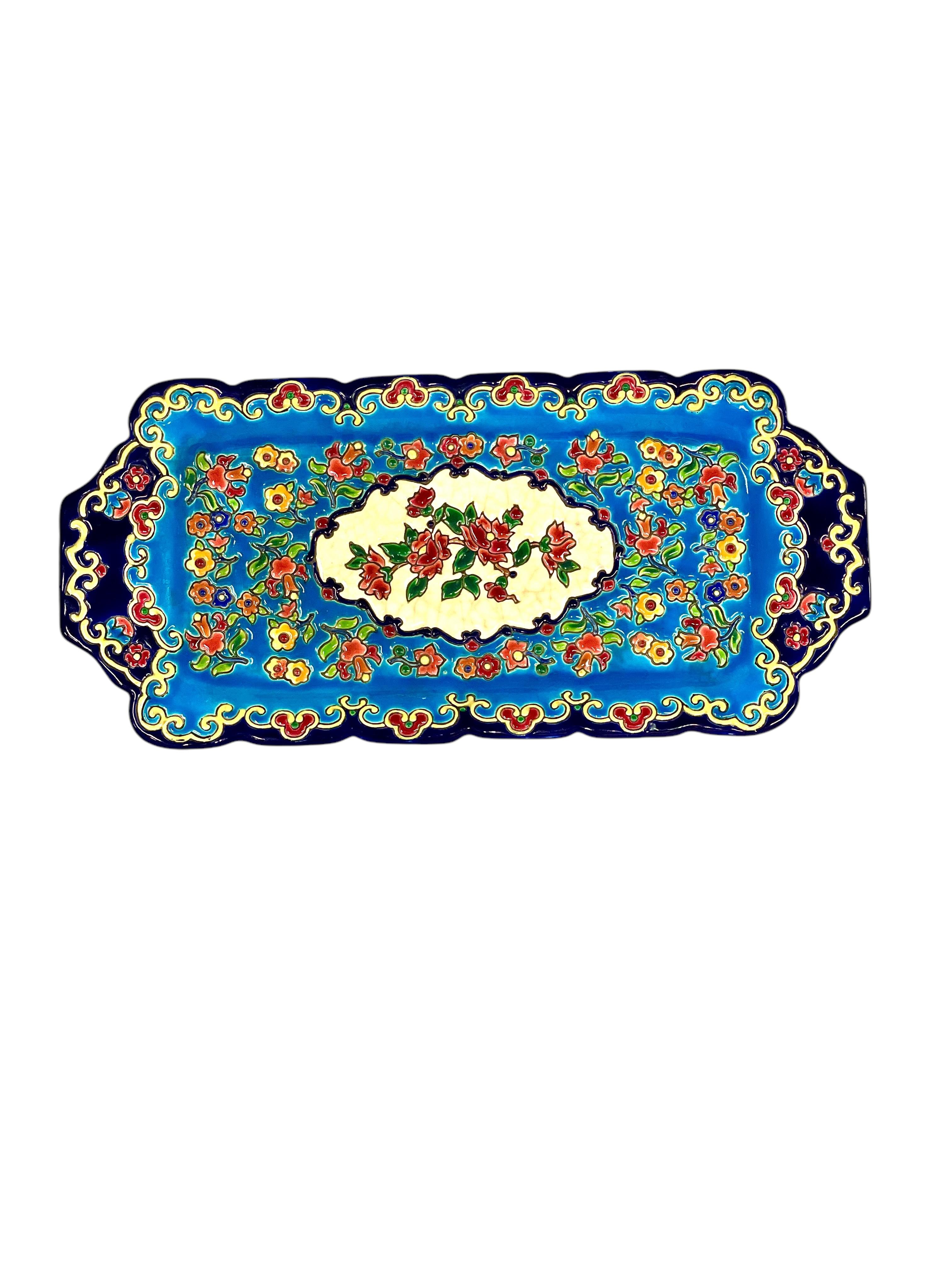 Esta exquisita fuente rectangular de cerámica es un bello ejemplo de la artesanía tradicional francesa de Émaux de Longwy. Decorada a mano con vibrantes motivos florales, presenta una rica paleta de colores dominada por azules profundos, turquesas y