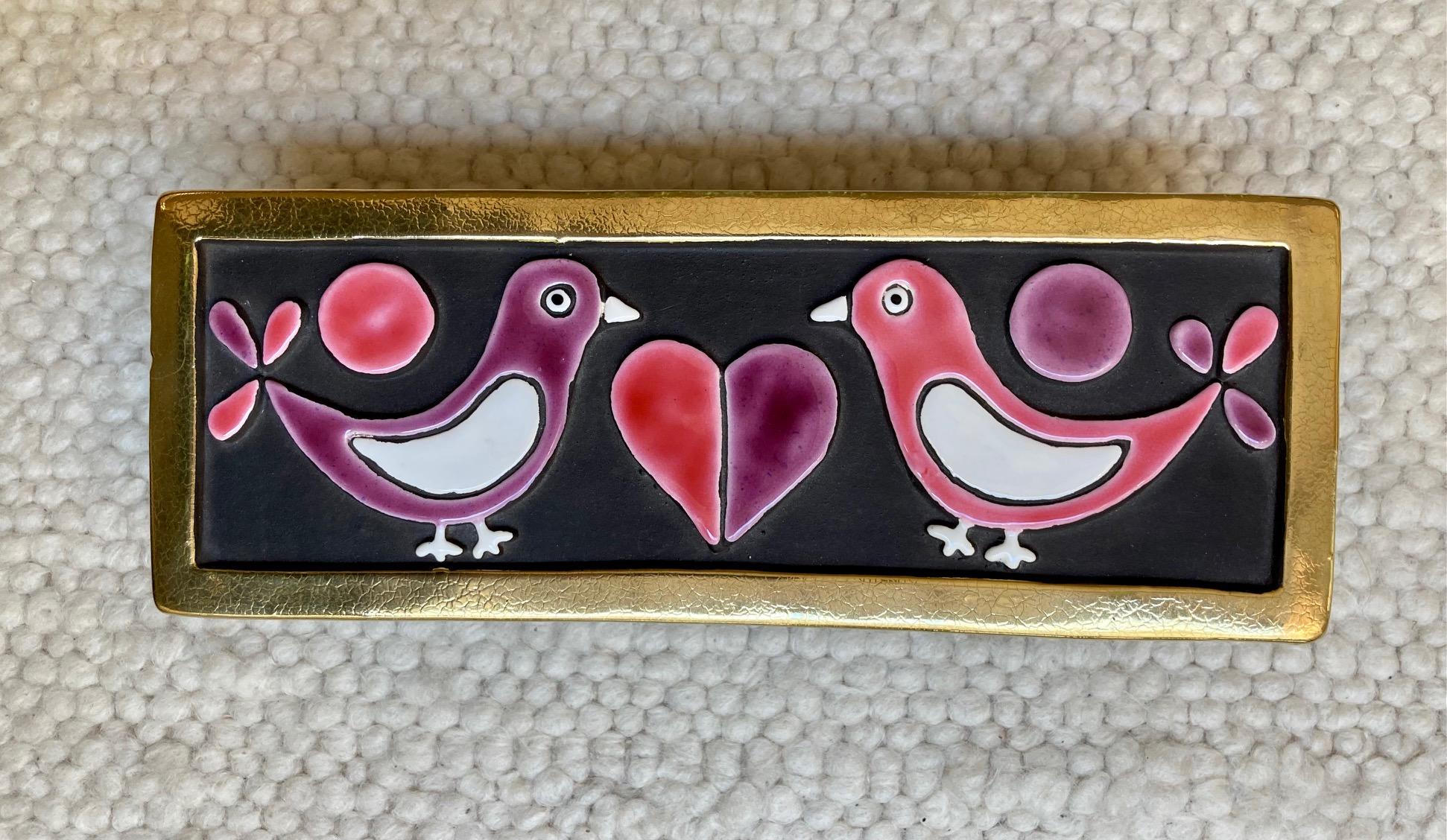 An enameled ceramic box sporting 2 doves facing each other and a heart in the middle.
Embossed earthenware enameled in pink, purple and white on a black background. 
Crackled gold edges. 
the base of the box is in wood
Model 