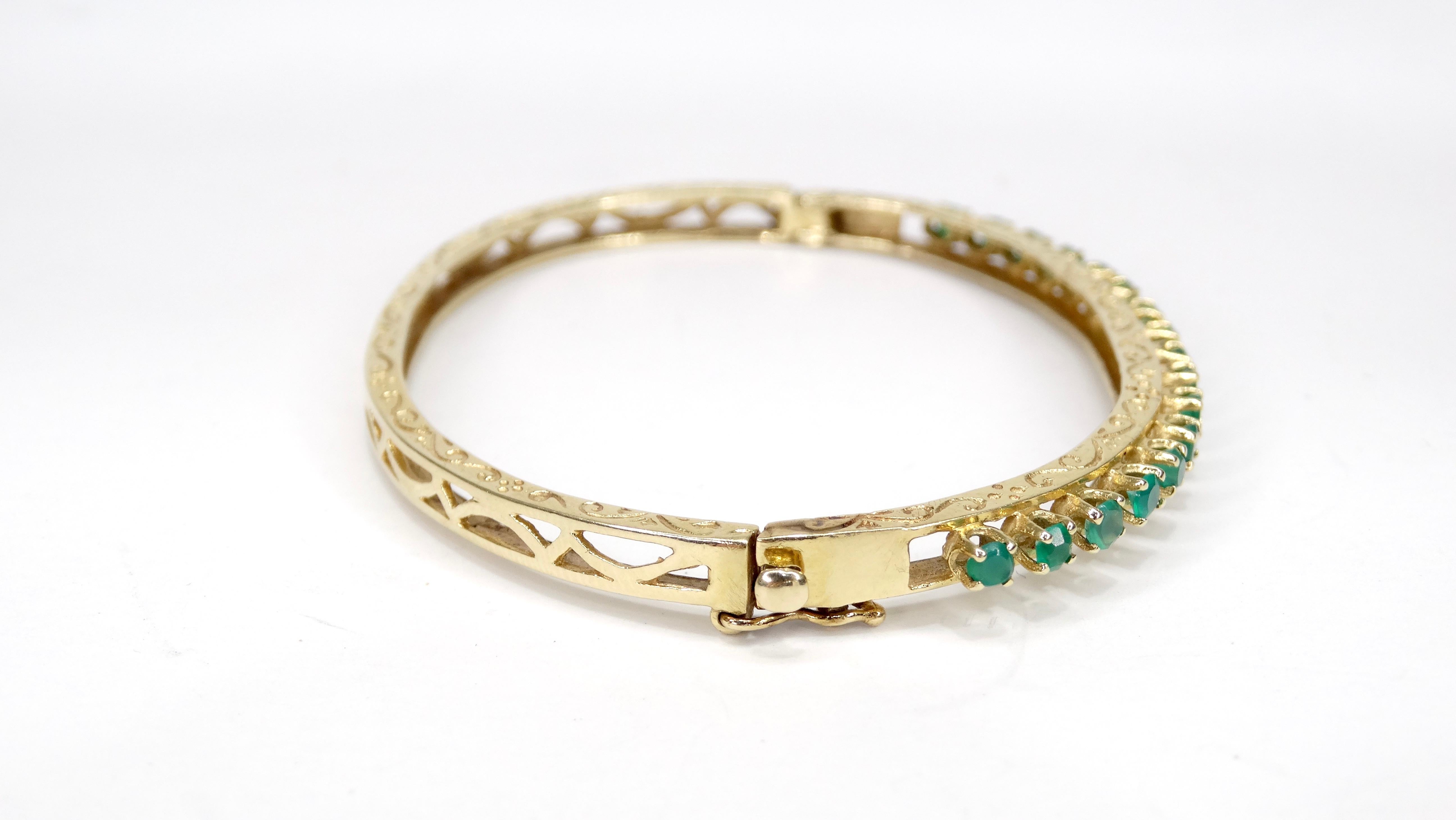 Circa mid 20th-century, this beautiful bangle is crafted 14k gold and features a row of round cut Emeralds in a prong setting. Bracelet is embossed with a decorative pattern and includes a press closure with a security clasp. Stamped 14k and weighs