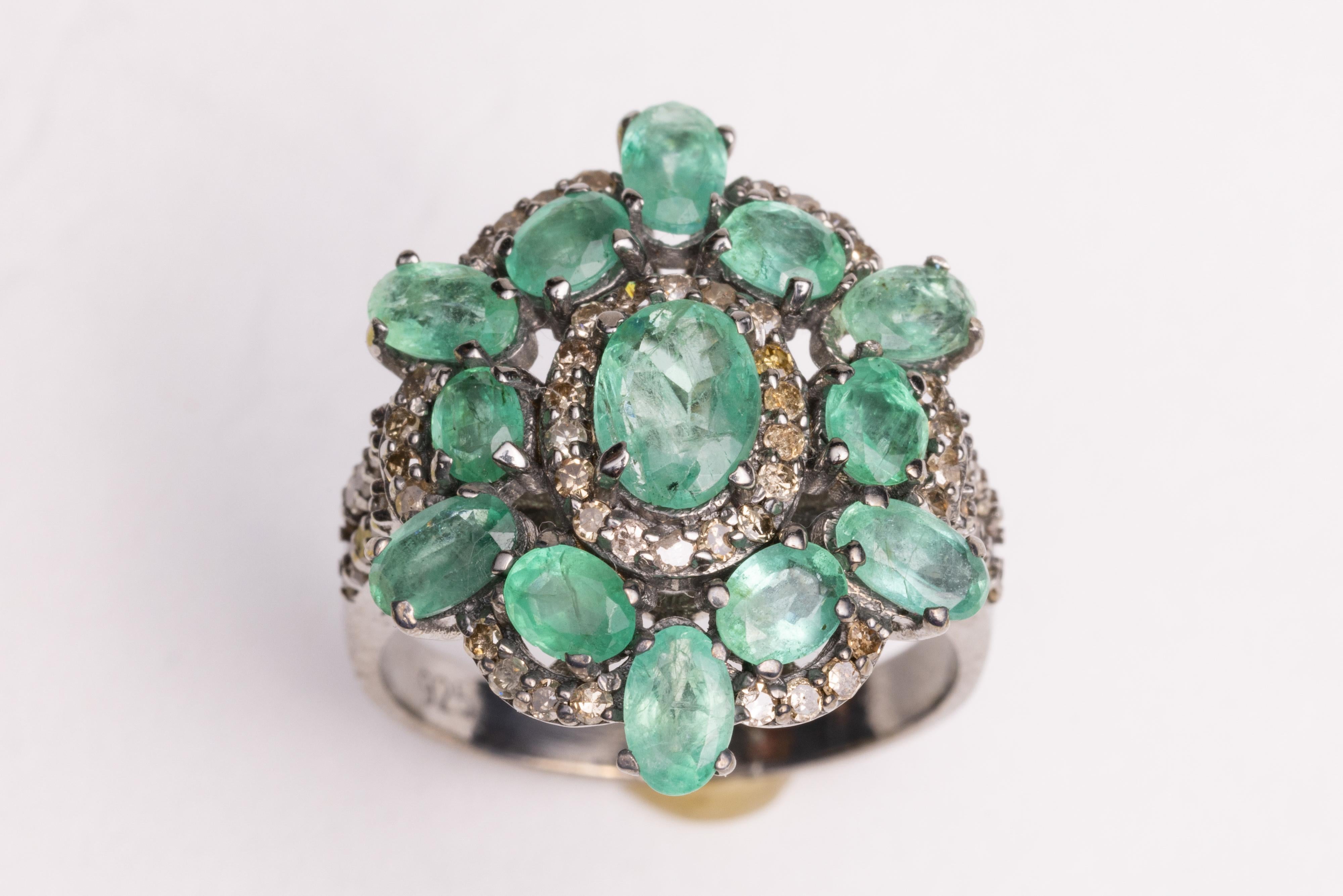 A fabulous cocktail flower ring with a large central oval, faceted emerald with 12 slightly smaller oval, faceted emeralds.  All are surrounded by pave`-set, round and brilliant cut diamonds, and diamonds on the double-band's shoulder as well.  Set