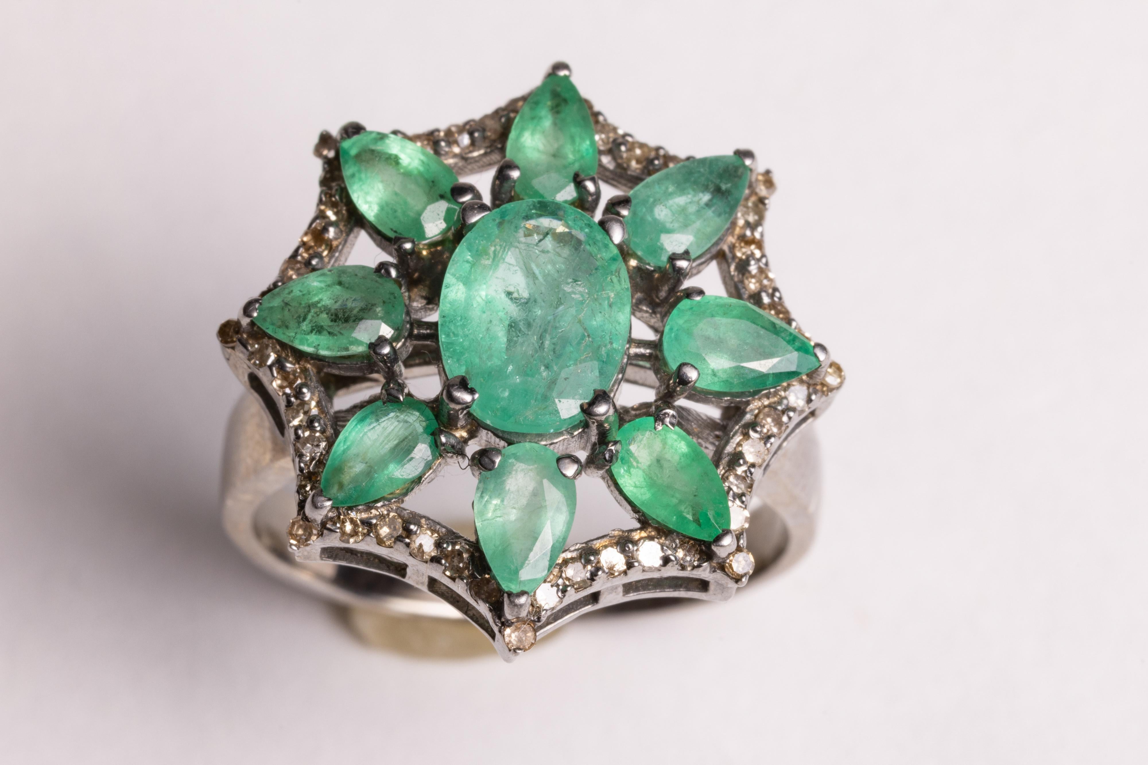 An emerald cluster cocktail ring with an oval, faceted emerald in a flower motif surrounded by 8 pear-shaped faceted emeralds and round, brilliant cut diamonds.  Set in sterling silver.  Carat weight of emerald is 2.42 carats, diamonds total .34