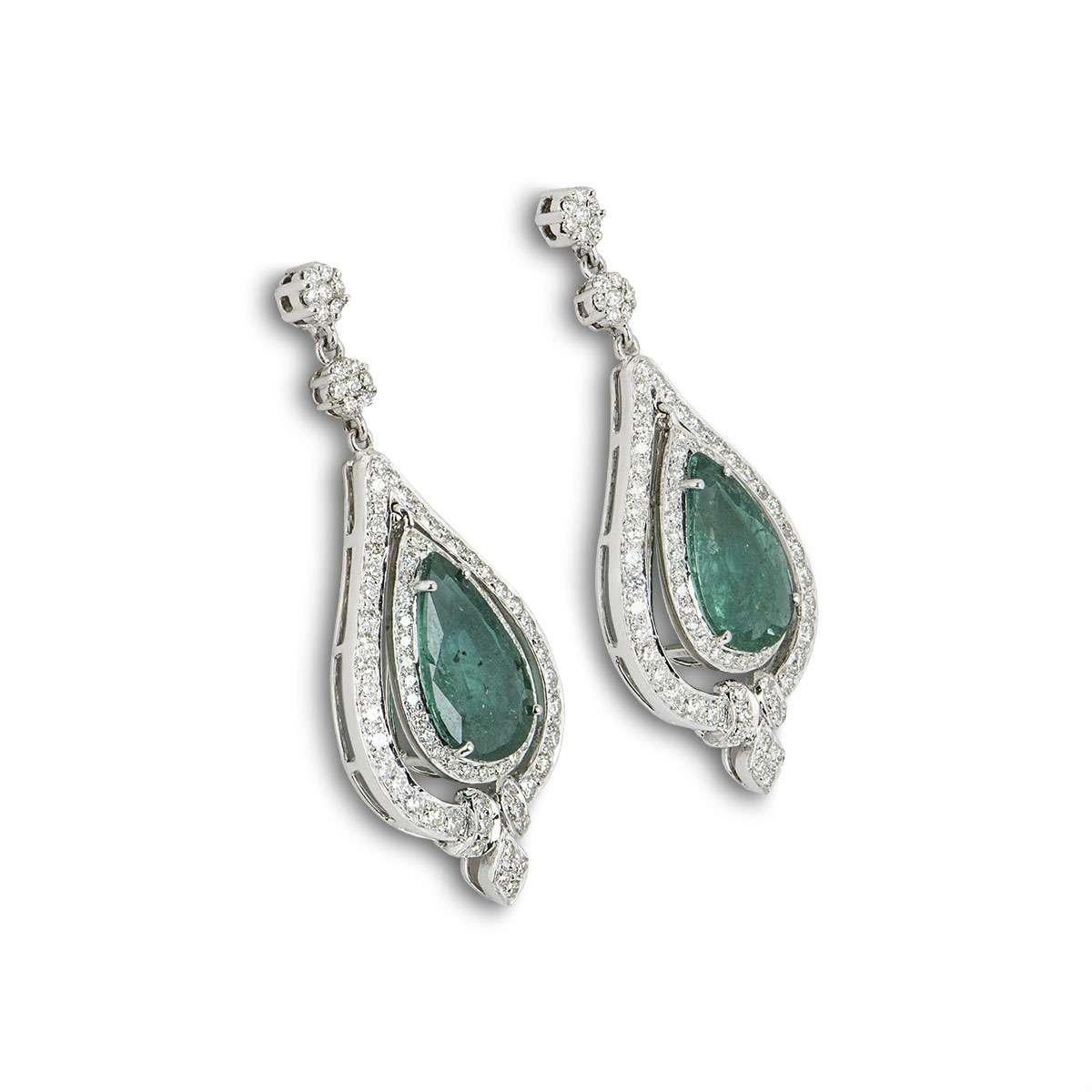 A pair of 18k white gold emerald and diamond drop earrings. The earrings are each set with a pear shaped emerald in the centre, surrounded by a double halo of round brilliant cut diamonds and two small diamond set flower motifs. The emeralds have a