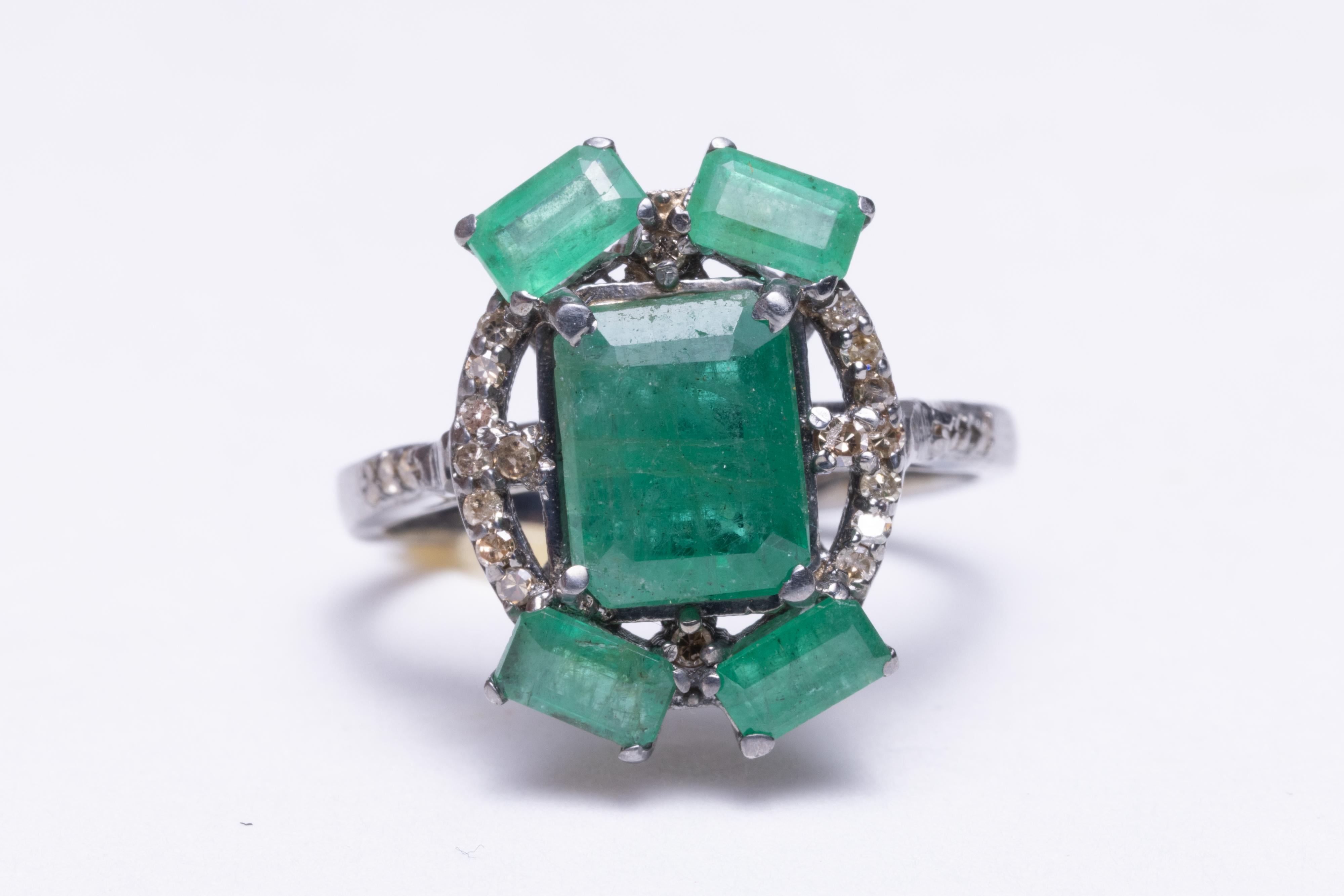 A large center emerald-cut emerald ring flanked with 4 smaller emeralds on the corners.  Pave`-set, round faceted diamonds around the face and along the shoulders of the band.  Set in an oxidized sterling silver.  Diamonds total .22 carats and the