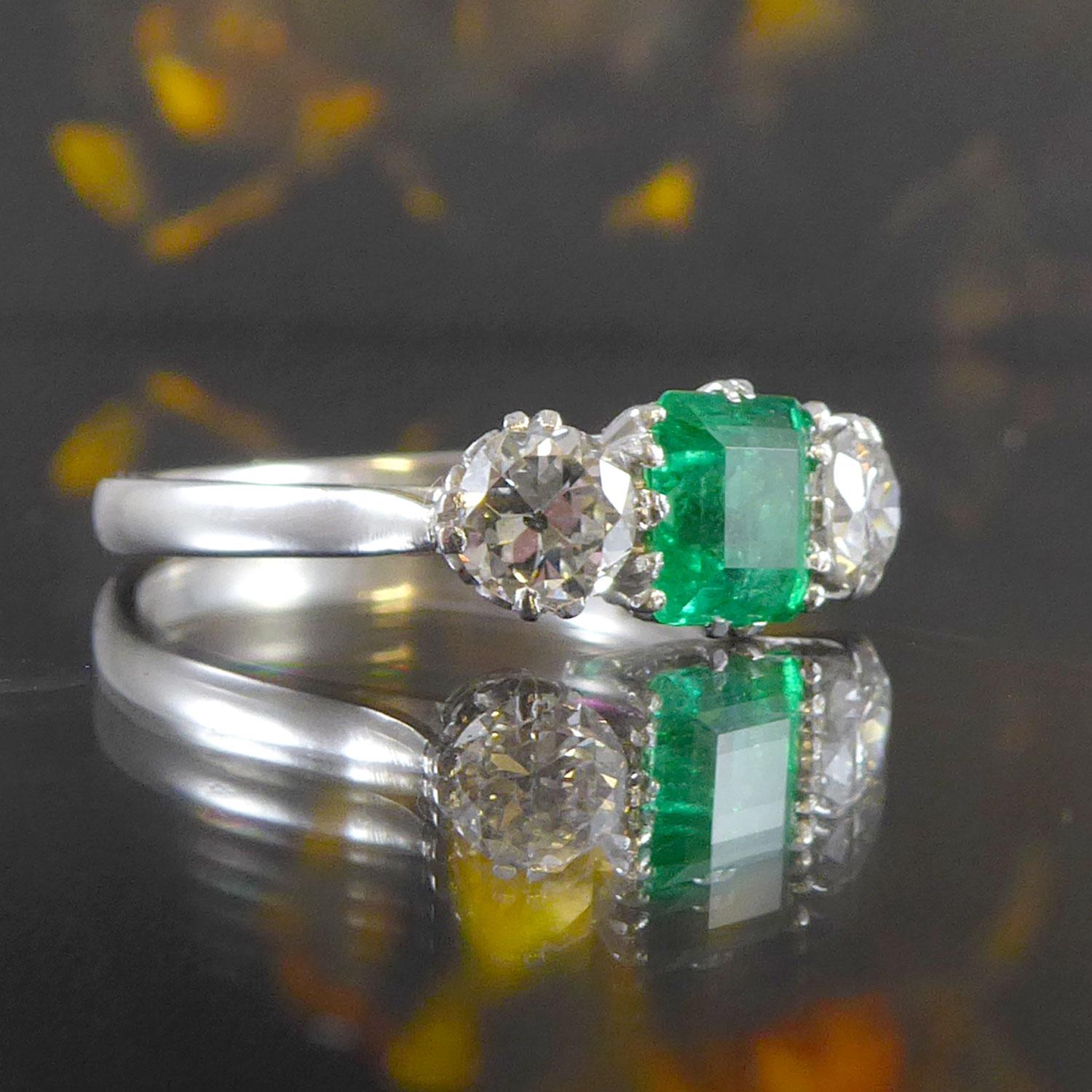 An emerald and diamond set three stone ring set with a rectangular shaped step cut emerald approx. 5.88mm x 4.66mm x 3.82mm deep and set within a double six claw basket style setting.  The emerald is flanked to each side by an early brilliant cut
