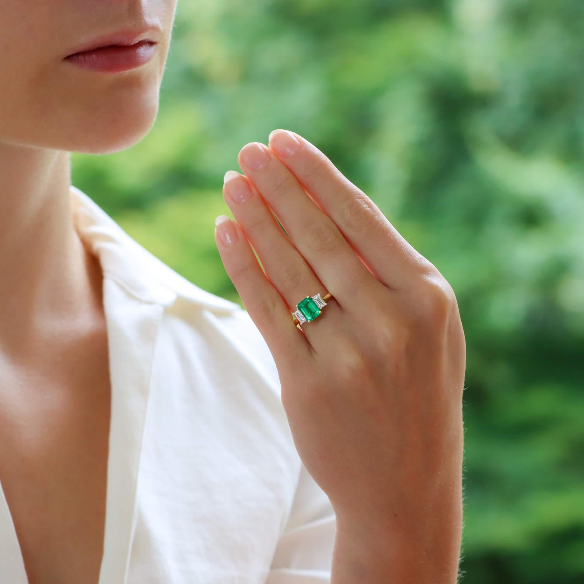 An elegant emerald and diamond three stone engagement ring set in 18k yellow and white gold. 
The ring is centrally set with an emerald cut emerald of vibrant green colour. It is four-claw set in yellow gold and sided with two perfectly matched