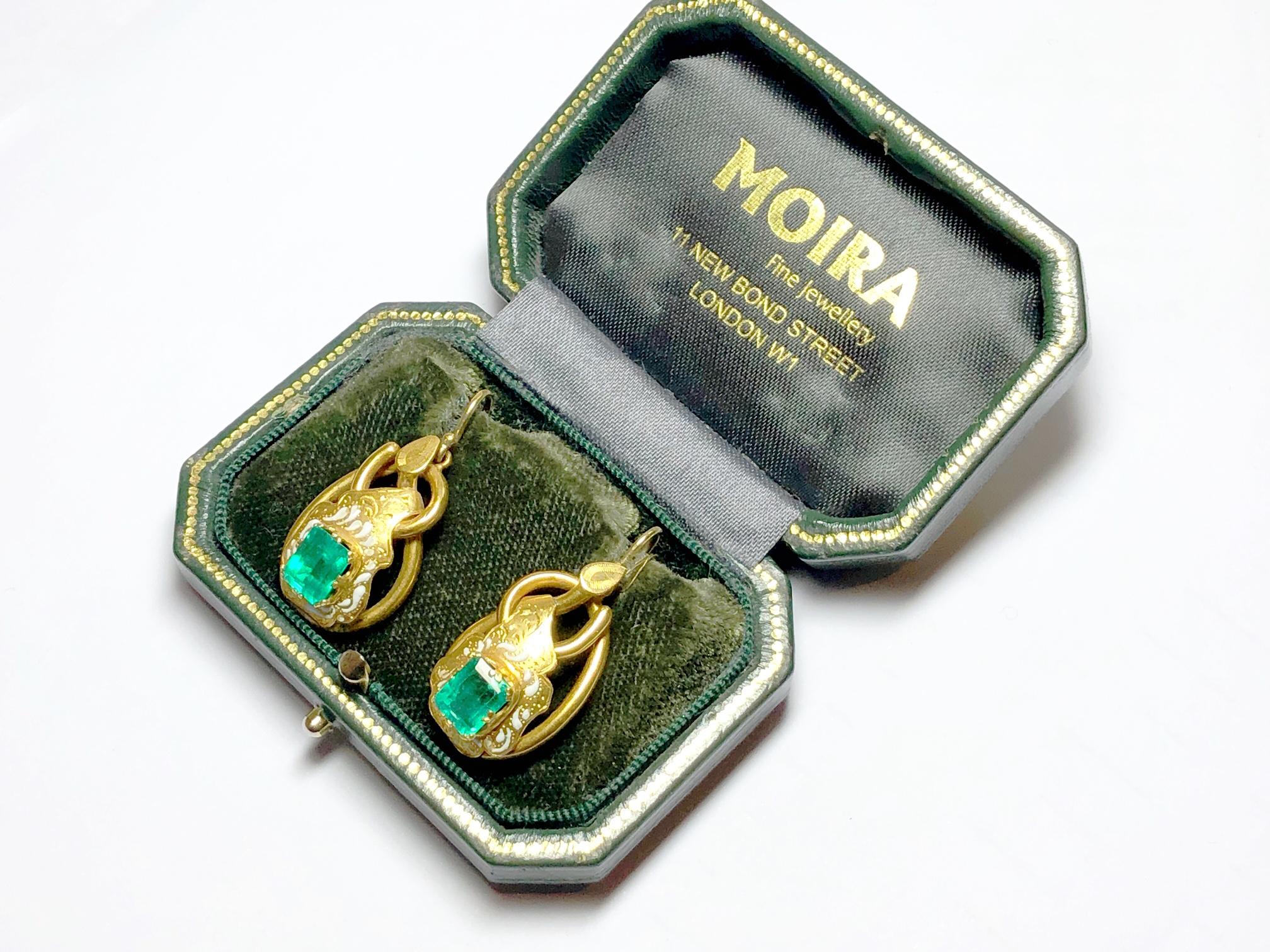 A pair of antique emerald and gold earrings, with a central emerald-cut emerald, weighing an estimated 1.40ct each, surrounded by an ornate engraved panel with white enamel detail, mounted on a gold double loop, French wire fittings. Circa 1880.