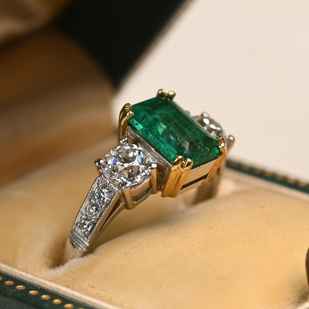 This elegant ring features a striking emerald-cut emerald weighing approximately 1.00 carat, secured in rich 18k yellow gold prongs. The vivid green center stone is flanked by two old European cut diamonds, creating a beautiful balance of shape and