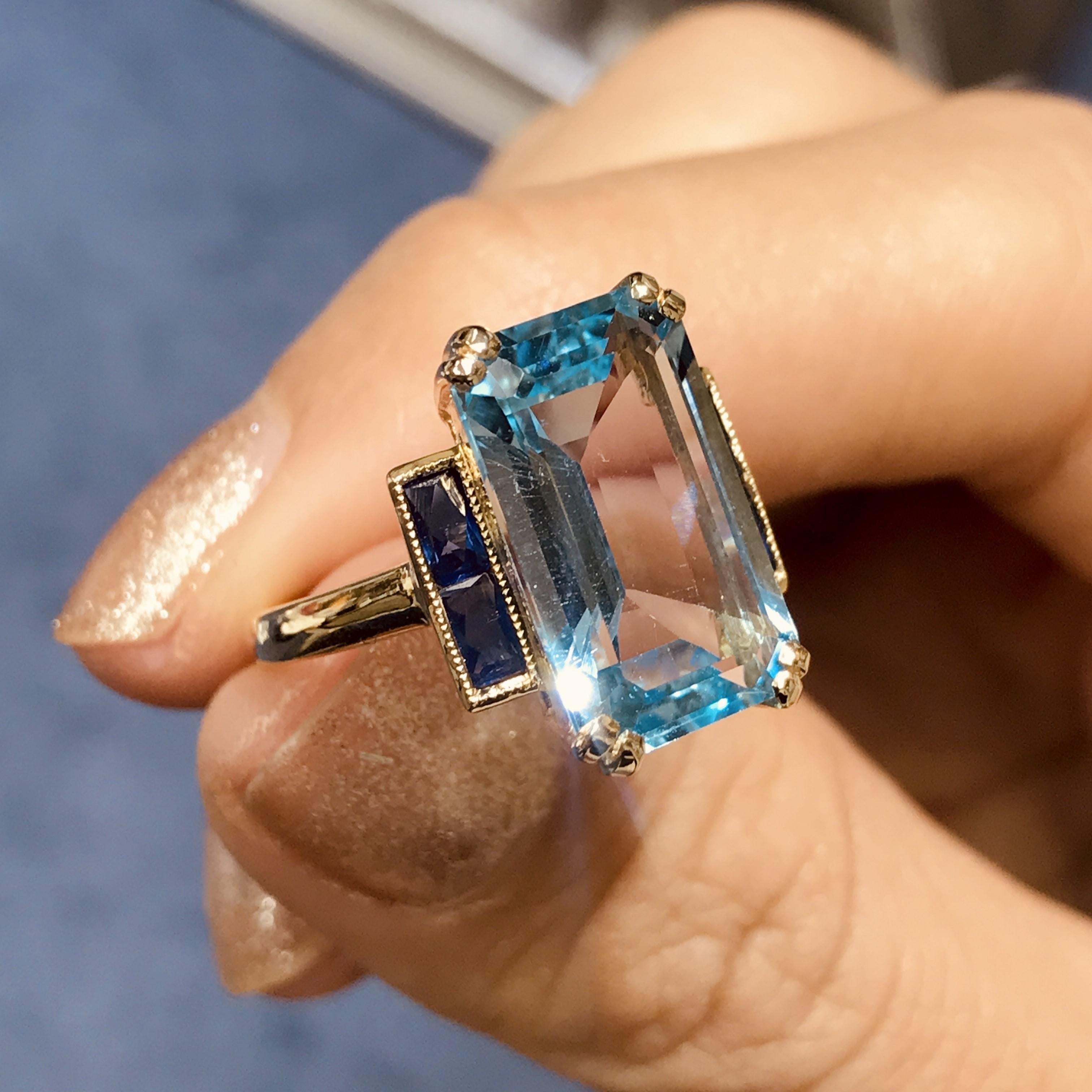 Questo elegante anello in stile Art Déco presenta al centro un topazio blu dal taglio smeraldo che irradia una brillantezza fresca e serena. La pietra centrale è affiancata su entrambi i lati da zaffiri blu intenso, che aggiungono contrasto e un