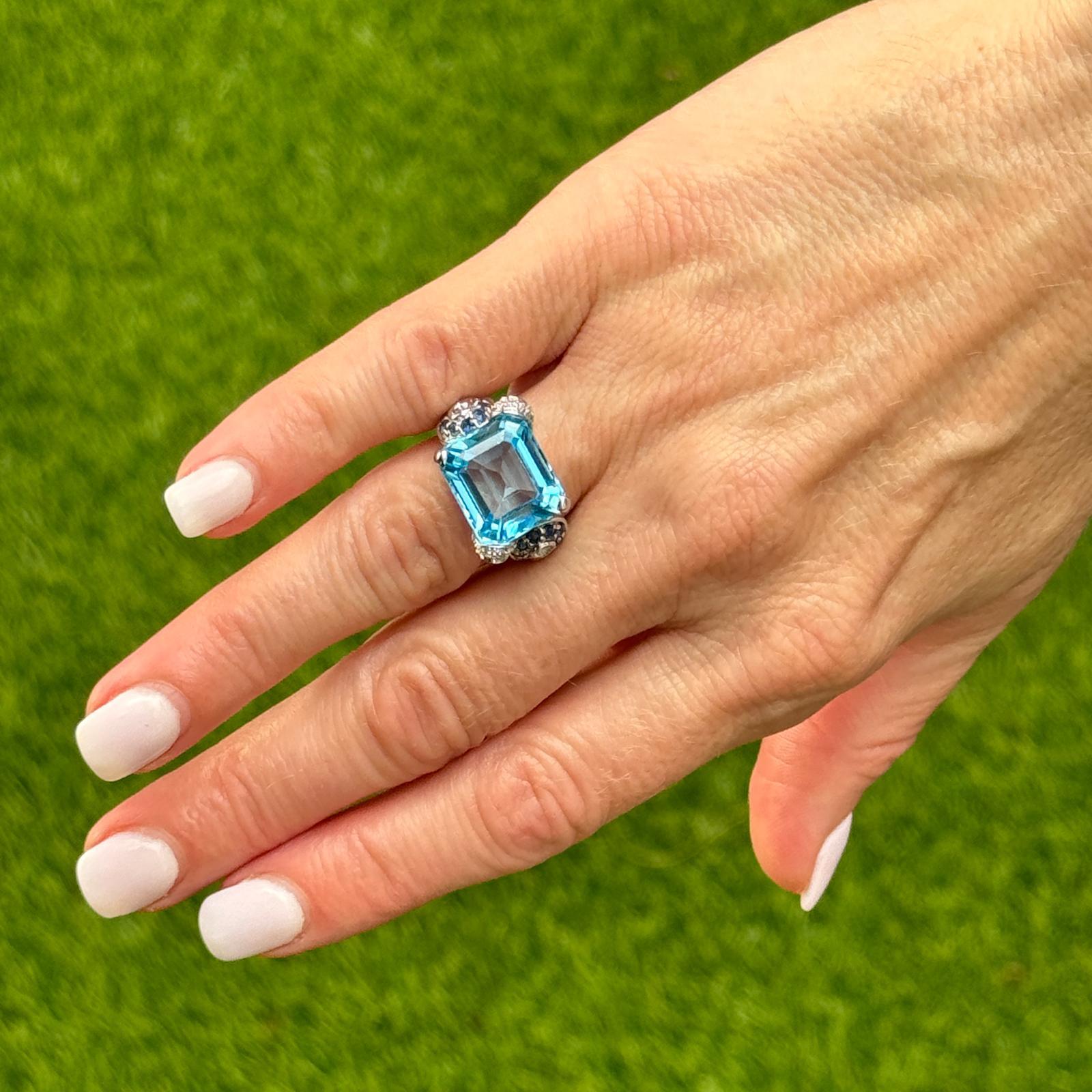 Modern blue topaz, sapphire, and diamond ring crafted in 14 karat white gold. The ring features an emerald cut blue topaz weighing approximately approximately 8.0 carats. The topaz is set east-west and the mounting is set with blue sapphire and