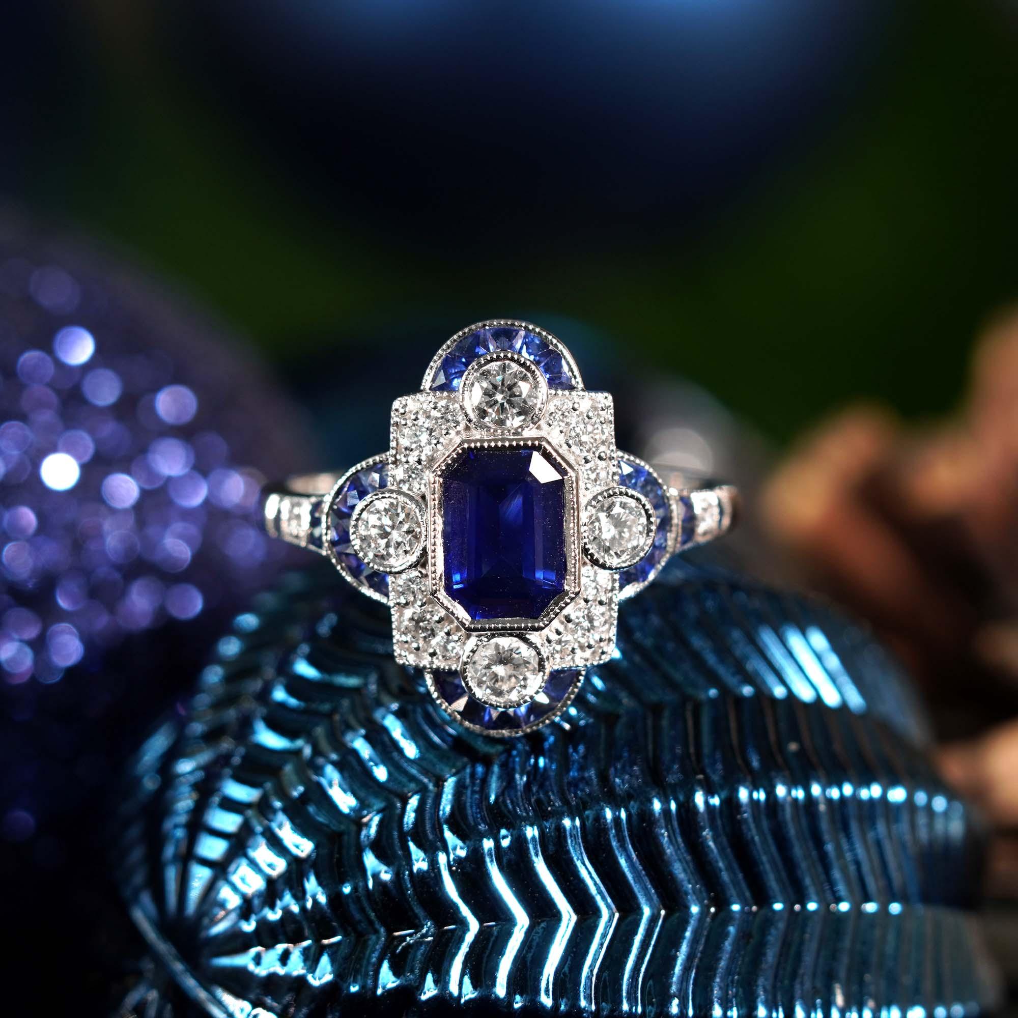 A striking and intriguing jewel, indeed. This bold Art Deco style ring highlights a gorgeous gleaming emerald cut Ceylon sapphire framed in eye-catching by white diamonds and French cut blue sapphires. Each side and shoulders set off with round