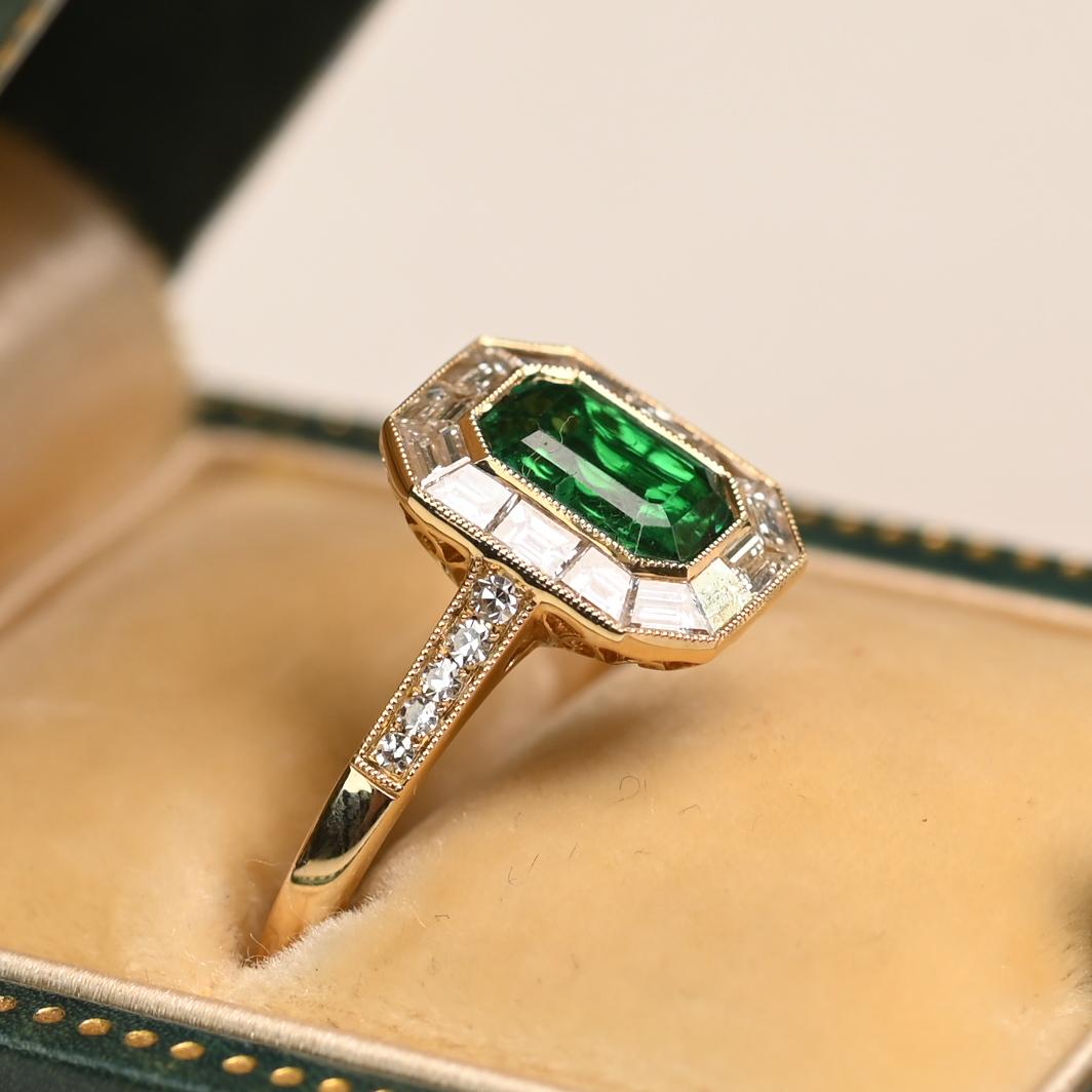 Im Mittelpunkt dieses eleganten Rings steht ein ca. 0,80 Karat schwerer Smaragd im Smaragdschliff, der sicher in einer Zargenfassung gefasst ist. Der leuchtend grüne Edelstein ist von einem raffinierten Halo aus kanalgefassten Baguette-Diamanten