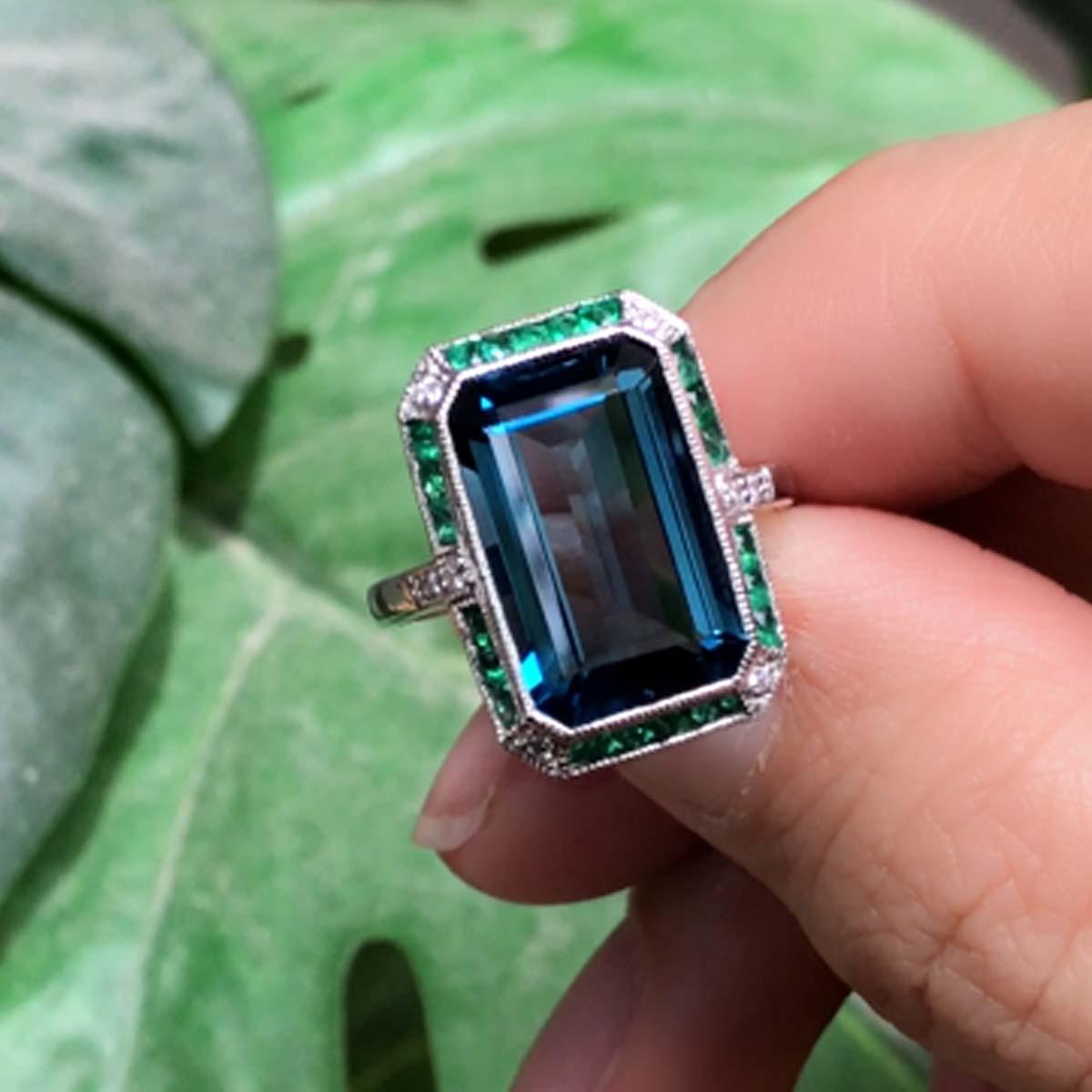 An Art Deco inspired 18K White Gold London blue topaz with emerald and diamond ring. The ring consists of one central emerald cut London blue topaz surrounded by French cut emeralds and round cut diamonds. 

Ring Information
Style: Art-deco
Metal: