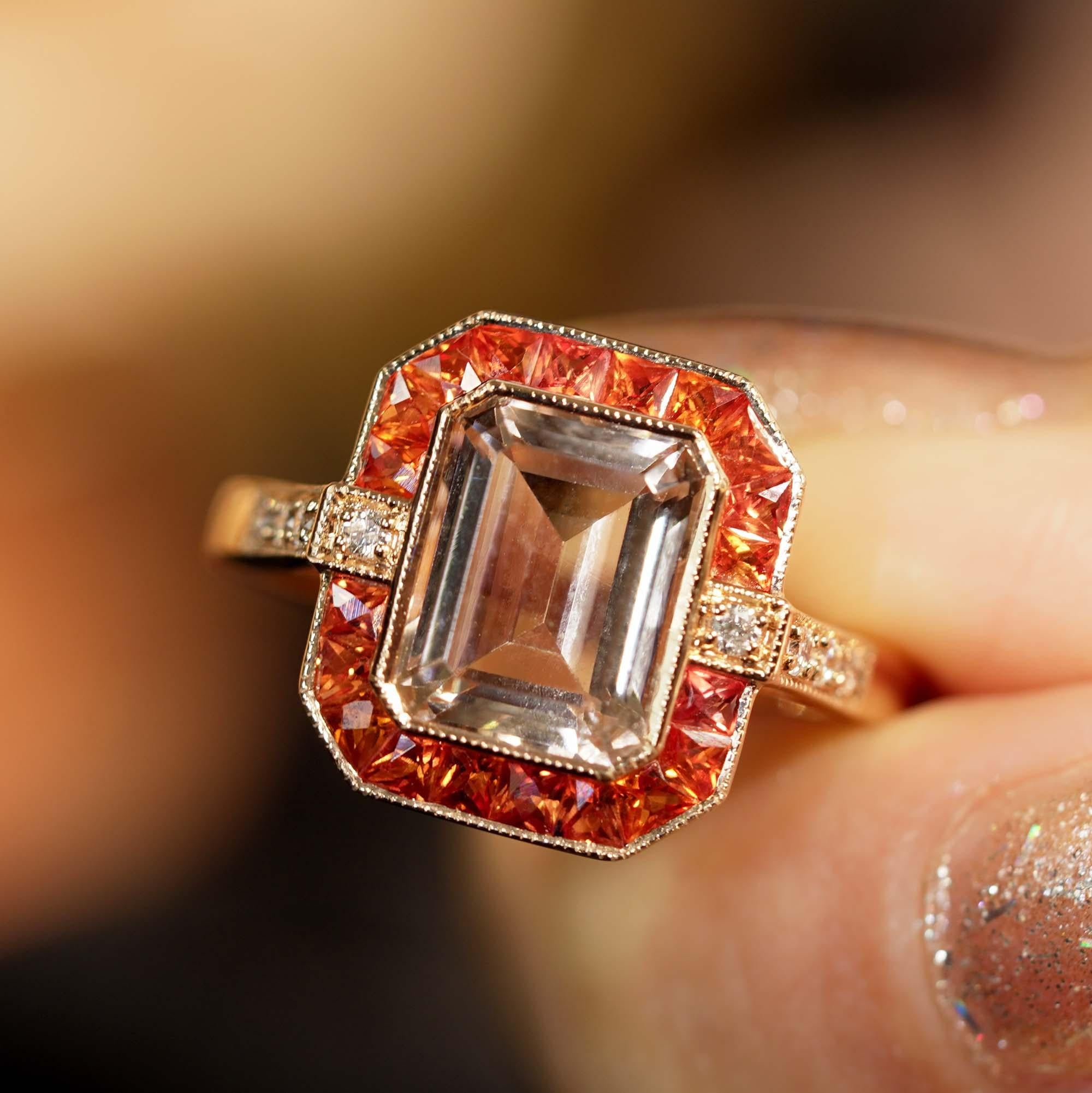 A striking and intriguing jewel, indeed. This bold and colorful Art Deco style ring highlights a gorgeous gleaming emerald cut 2.25 carat morganite framed in eye-catching color by French cut orange sapphires. Each side and shoulders set off with