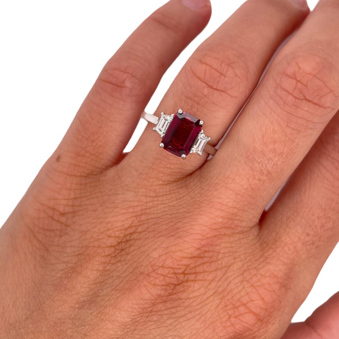 Classic three stone ruby and diamond ring in 18k white gold. Ring contains 1 center vivid red emerald cut ruby  2.07ct and 2 side step cut trapezoid diamonds 0.51tcw. Center ruby is BGL certifed with a Mozambique origin. Diamonds are G in color and