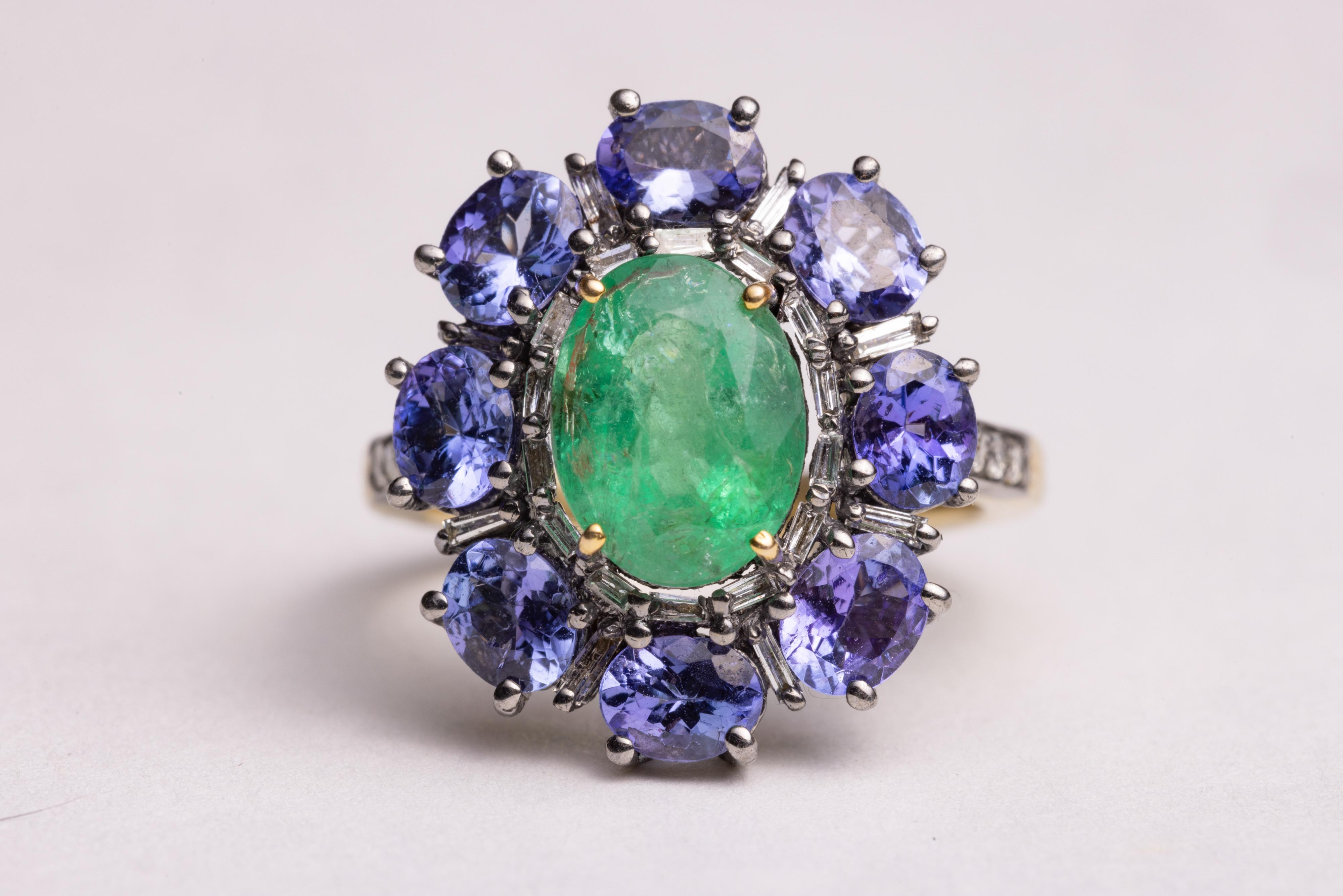 A cocktail ring with a large, oval faceted center emerald surrounded by baguette diamonds and oval faceted tanzanite gemstones.  Round brilliant cut diamonds along the side.  Set in sterling silver and vermeil.  Emerald stone is 2.75 carats, the