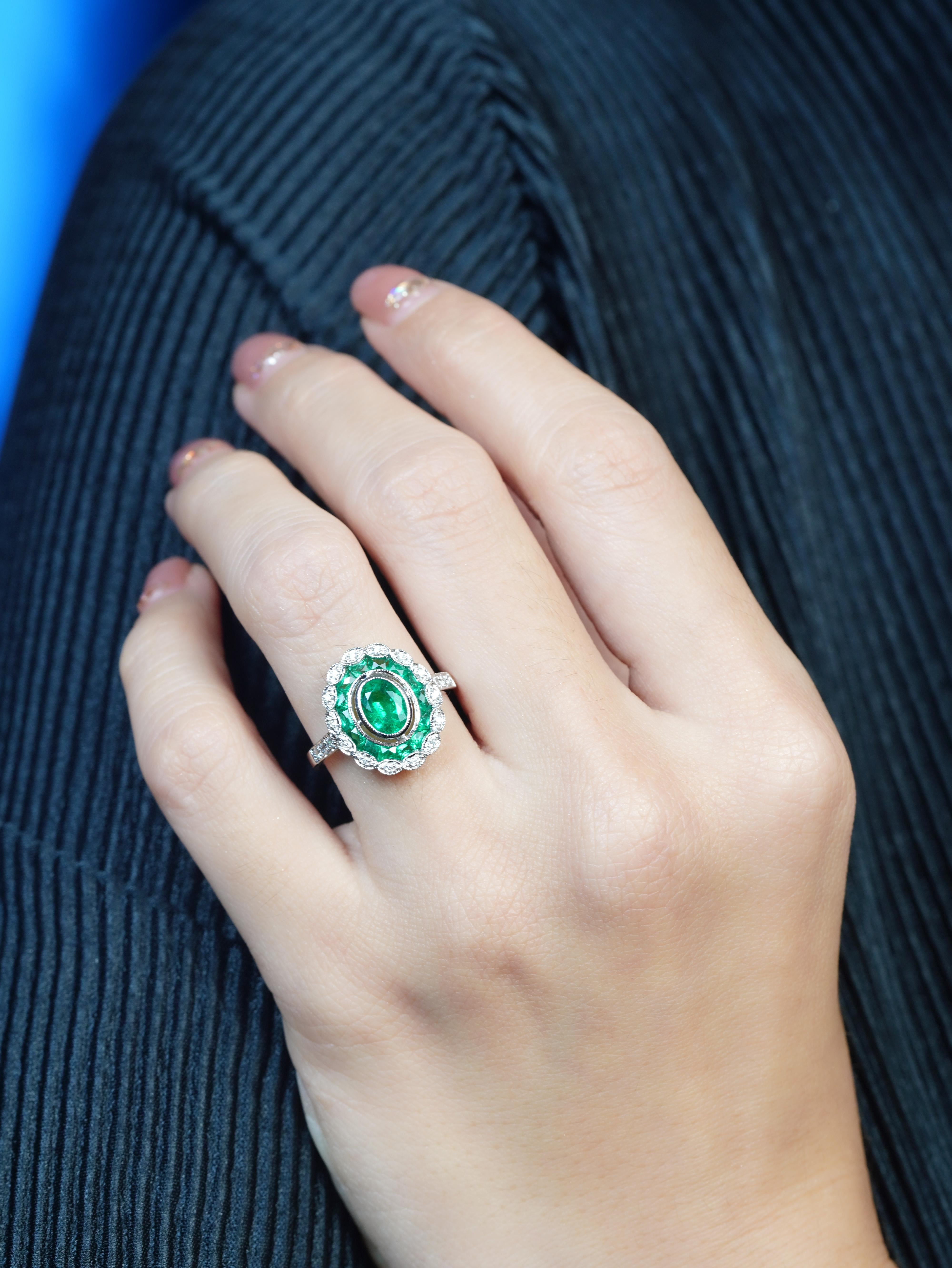 A bezel-set oval emerald, surrounded by a layer of French-cut emeralds that add depth and brilliance. The outer layer of diamonds evokes a delicate floral motif, blending geometric precision with nature-inspired elegance. Rich green tones contrast