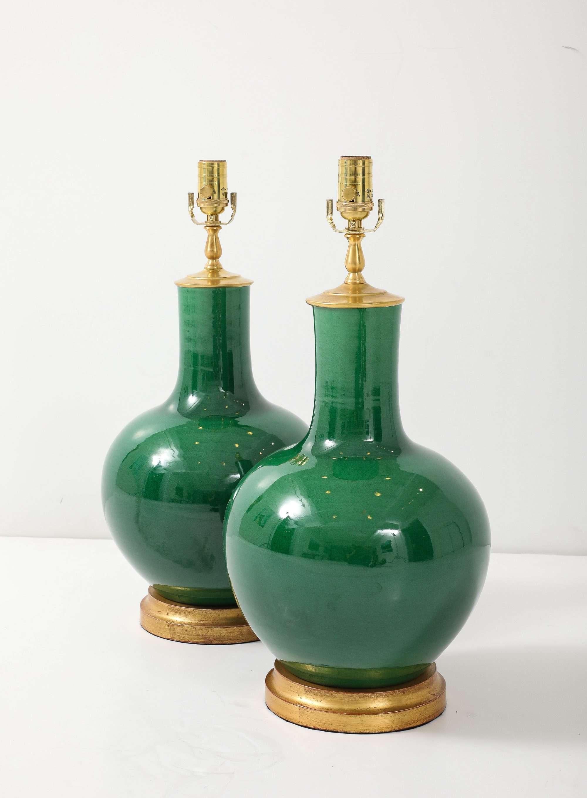 A pair of ceramic table lamps finished in a deep, high-gloss emerald glaze. Each lamp sits on a hand-gilded wood basand is capped with polished brass hardware, lending a tailored sparkle to the rich green body. The clean, sculptural silhouette