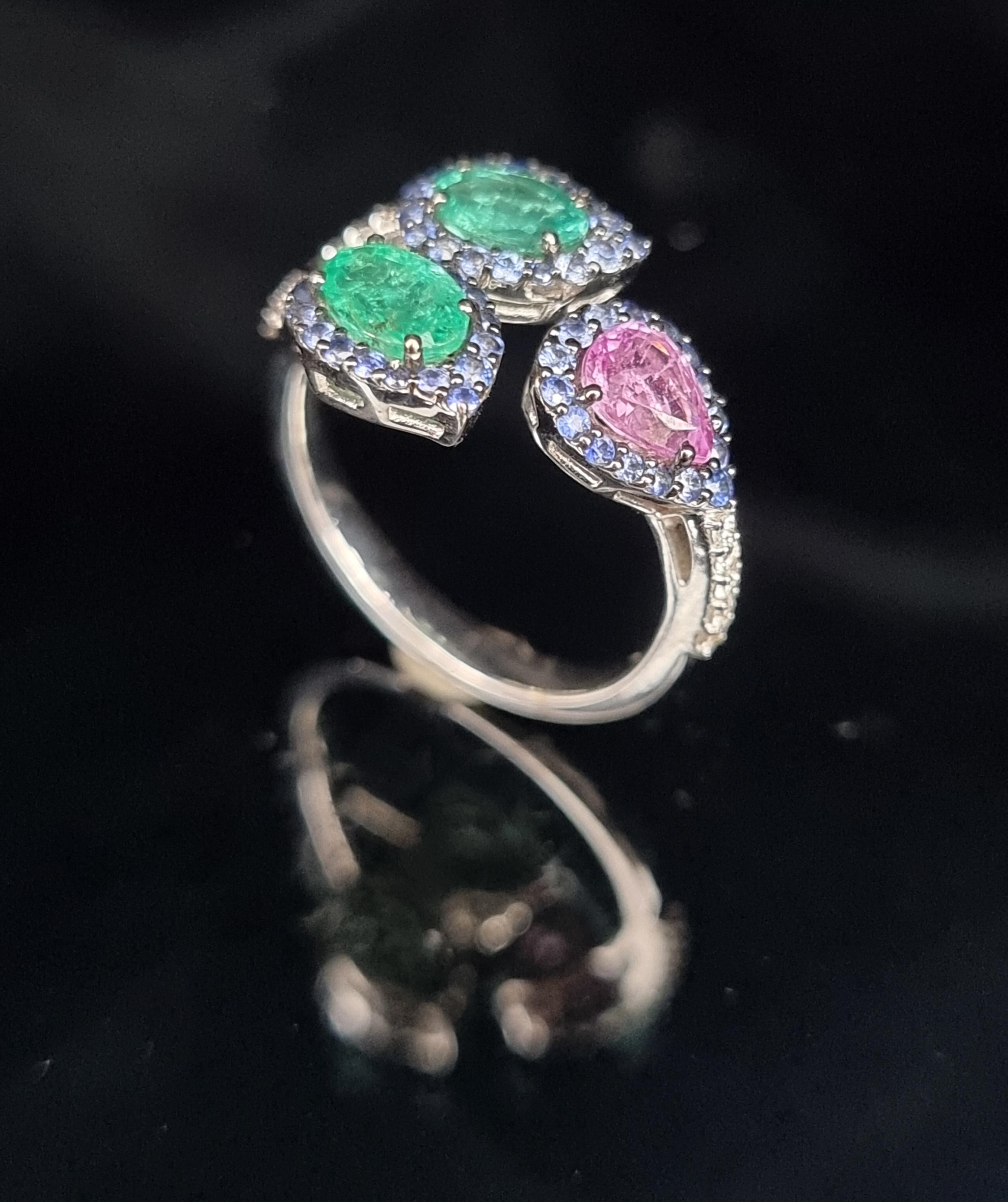 This stunning split ring is a masterpiece of elegance and contrast, featuring vibrant emeralds (0.99cts.) on one side and vivid pink sapphire (0.67cts.) on the other. Surrounding these captivating gemstones are a halo of deep blue sapphires