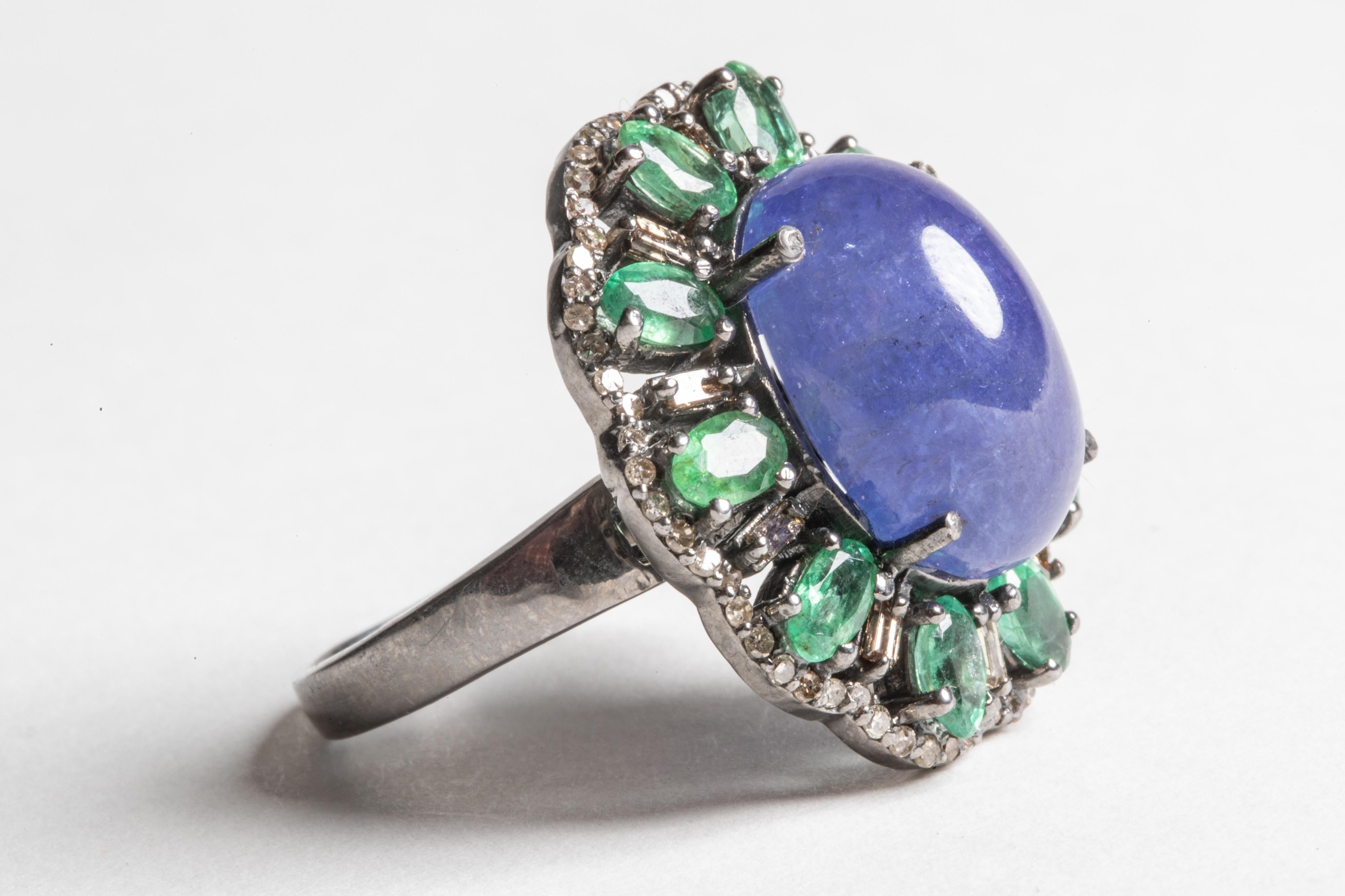 A large 14 carat cabochon Tanzanite dome ring surrounded by faceted, oval Columbian emeralds separated by baguette diamonds and round, brilliant cut diamonds all along the border.  Set in an oxidized sterling silver.  Ring size is 8.  Emeralds total