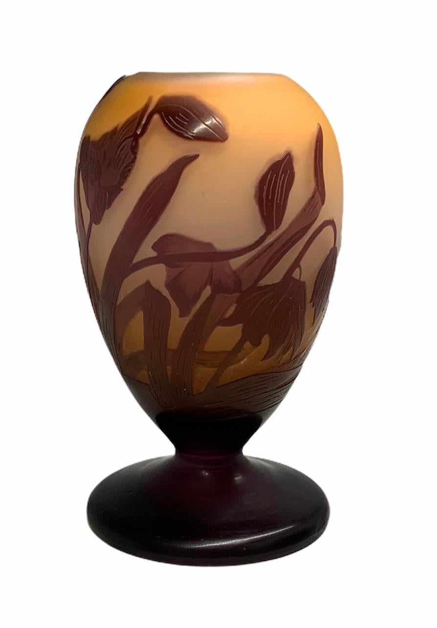 This is a Galle small urn shaped cameo vase. It has a frosted orange glass background highlighted by wine color branches of lilies flowers in movement. The wine color get to the round pedestal base and covers it completely. The glass vase is signed