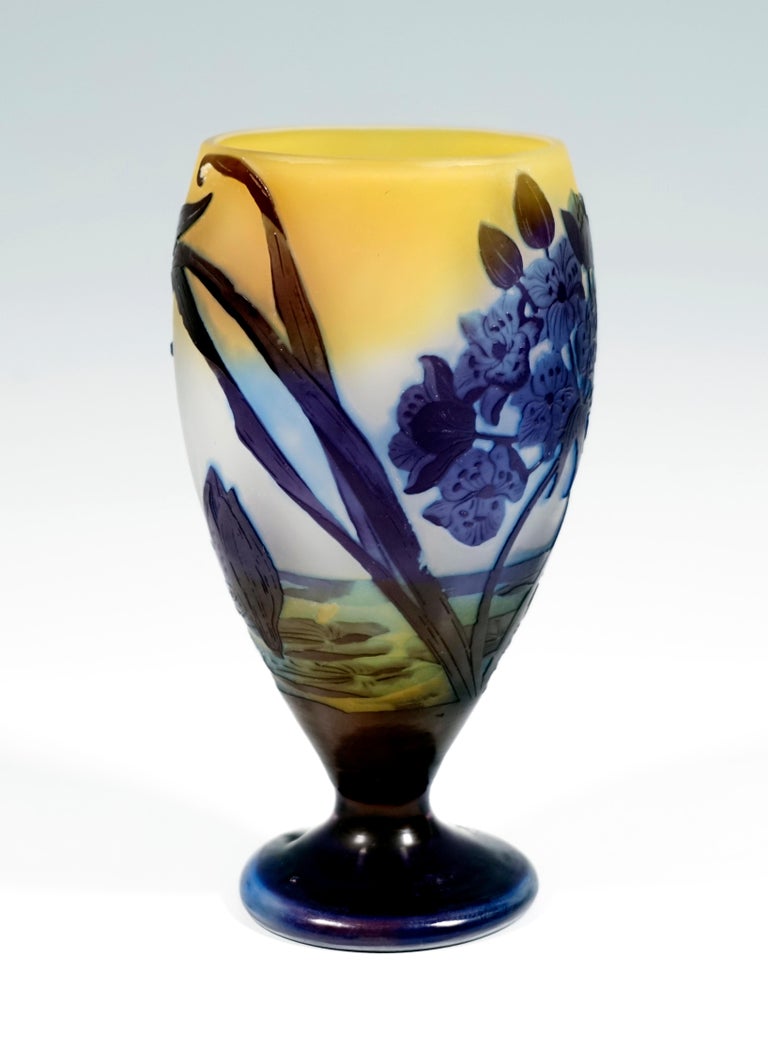 Vase in the shape of a cup: bulbous, tapering body, ending in a detached, round base. The area close to the ground is covered in dark blue overlay, above etched seascape decor with water lilies and flowers in front of a milky